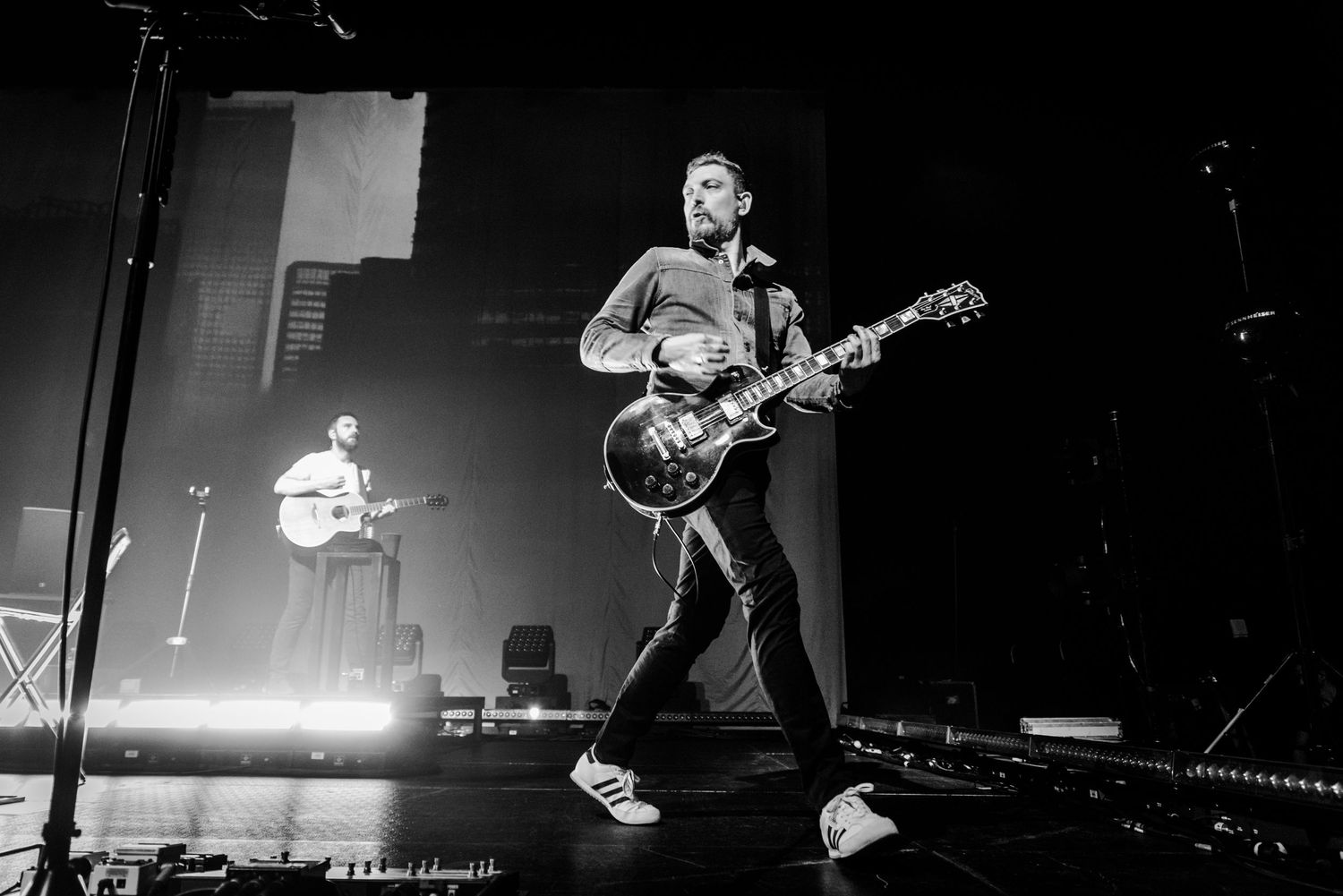 Black and white concert photo showing guitarist performing with stage lighting and second musician in background.