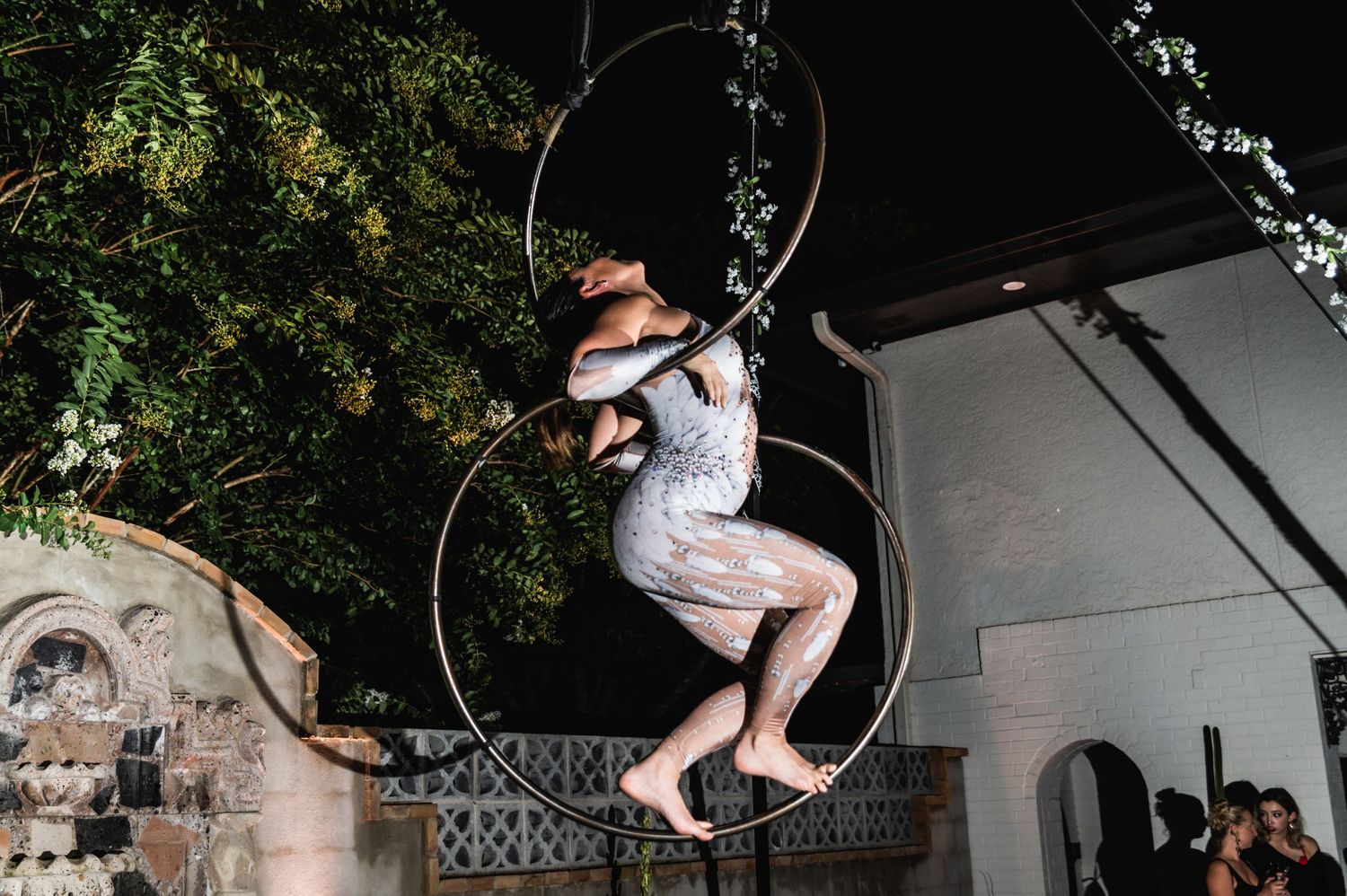 Aerial hoop performer in white costume against garden backdrop during evening show.