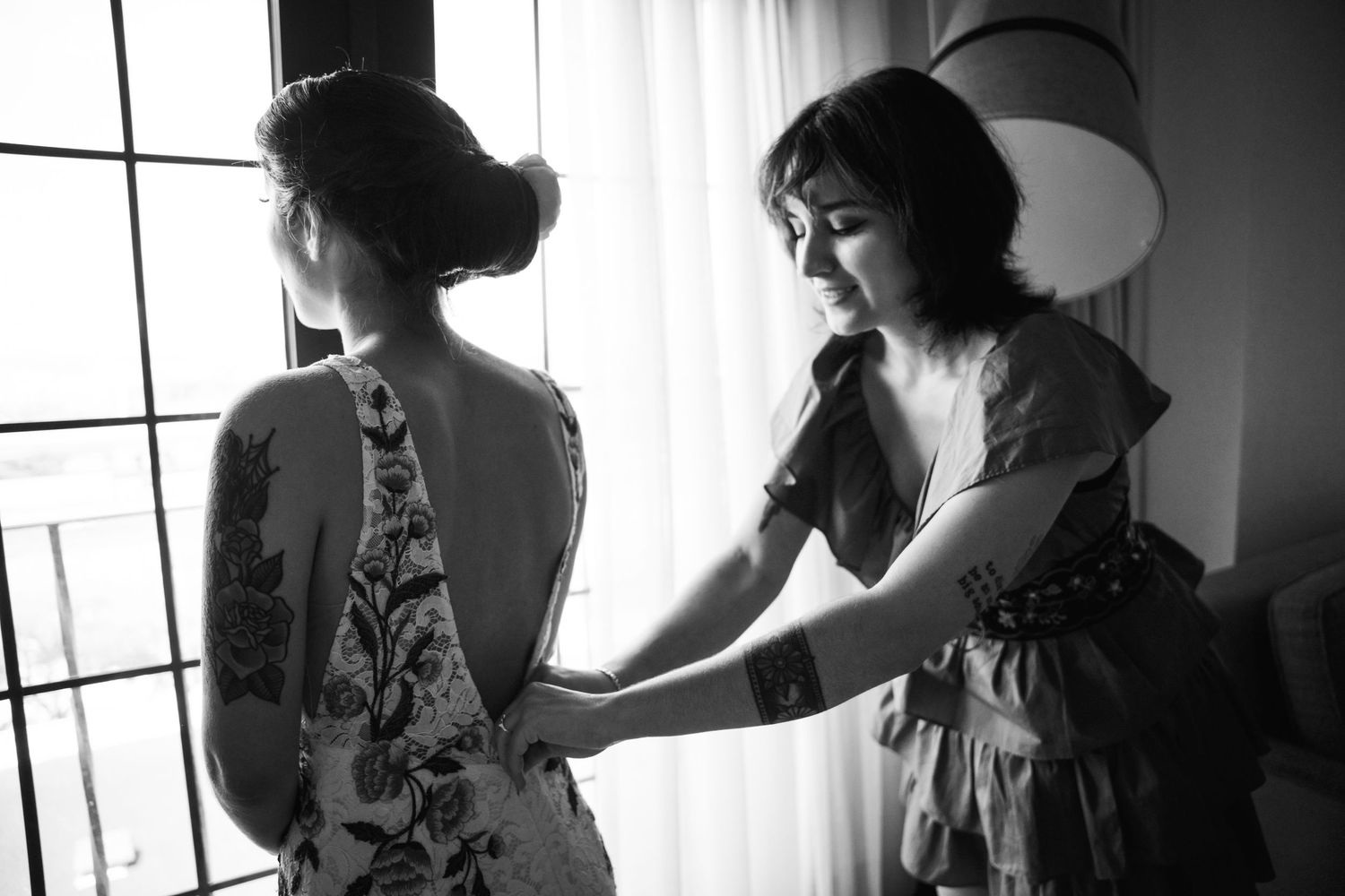 Two people in dresses prepare for an event near a window in black and white.