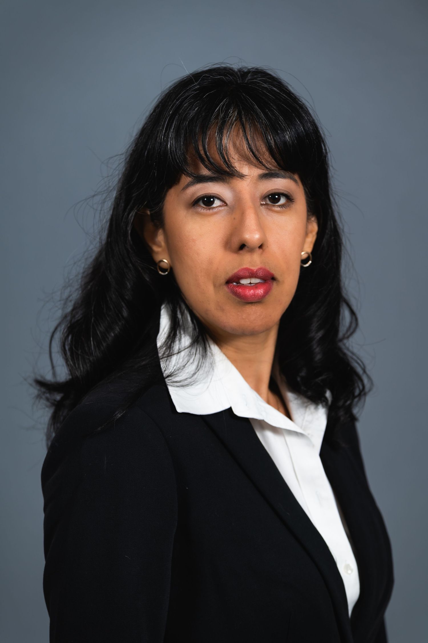 Professional headshot of a woman wearing a black blazer and white shirt against grey background.