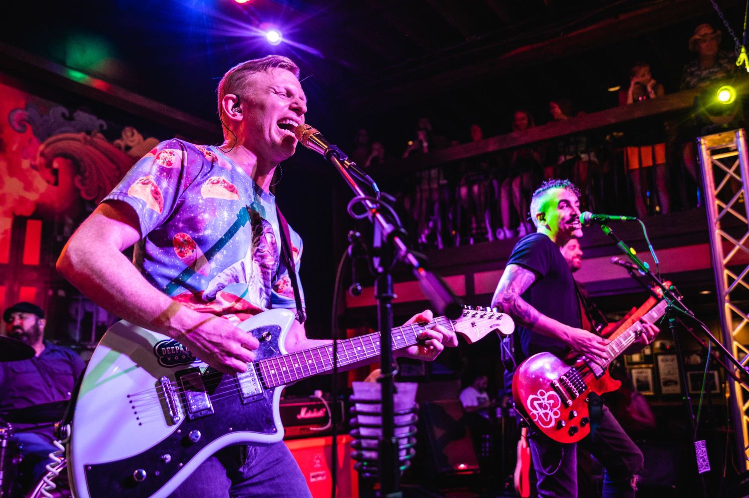 Musicians perform on stage with guitars under colorful lighting at an intimate venue.