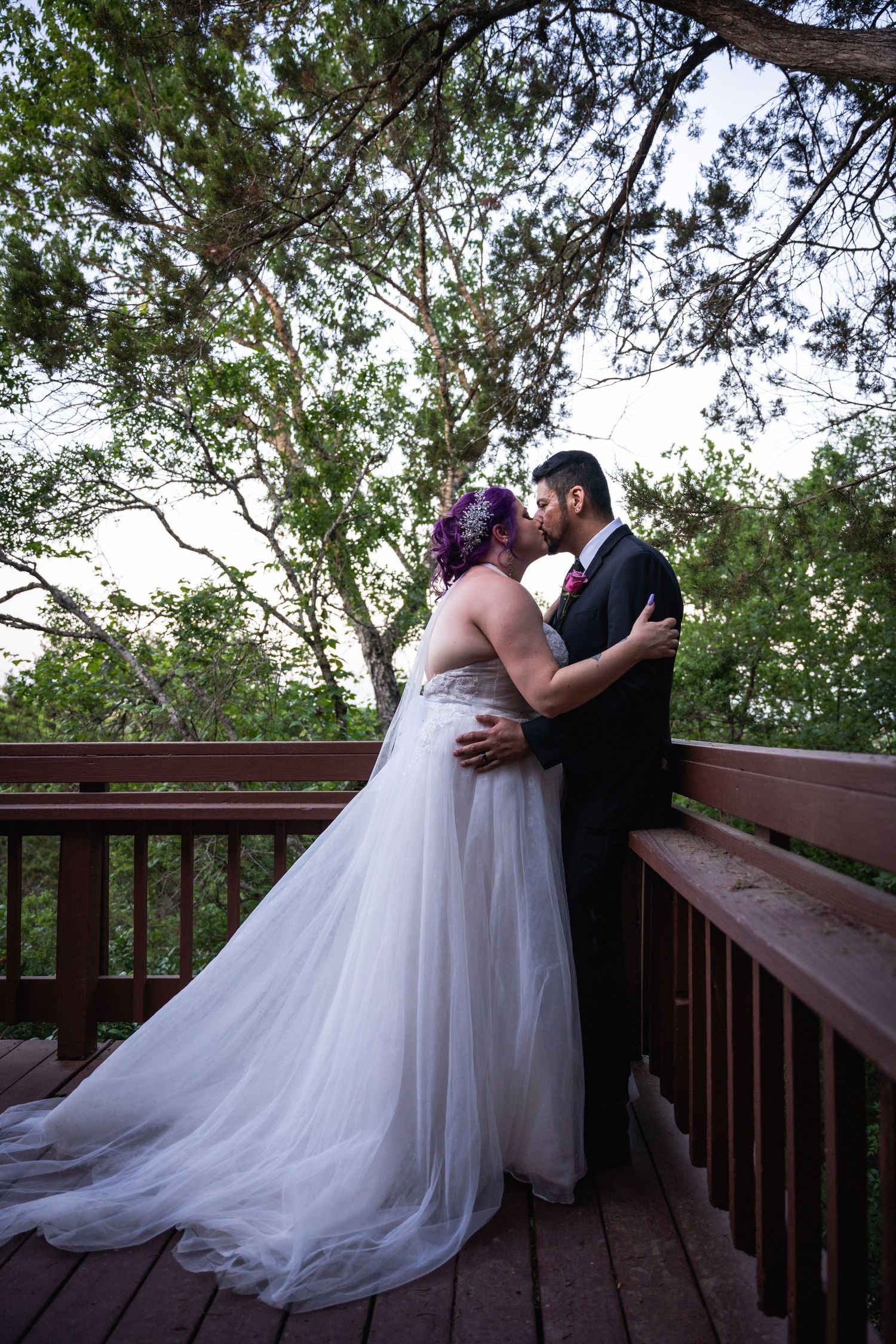A romantic embrace between newlyweds on a wooden deck surrounded by tall trees.