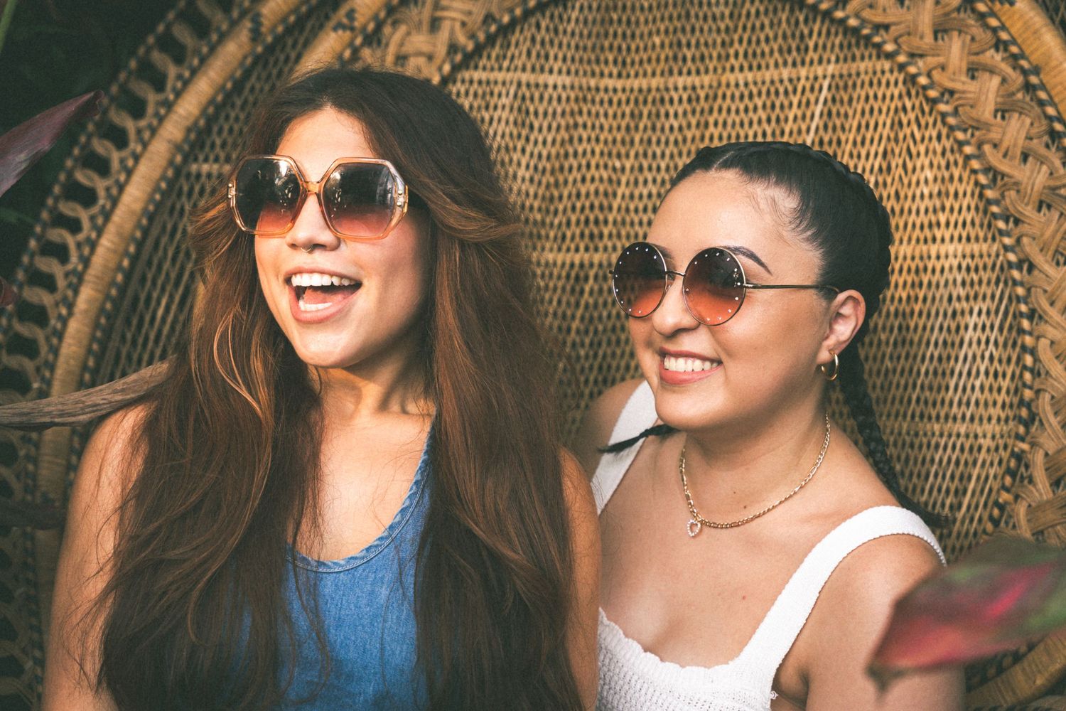 Two friends laughing while sitting on wicker furniture wearing sunglasses.