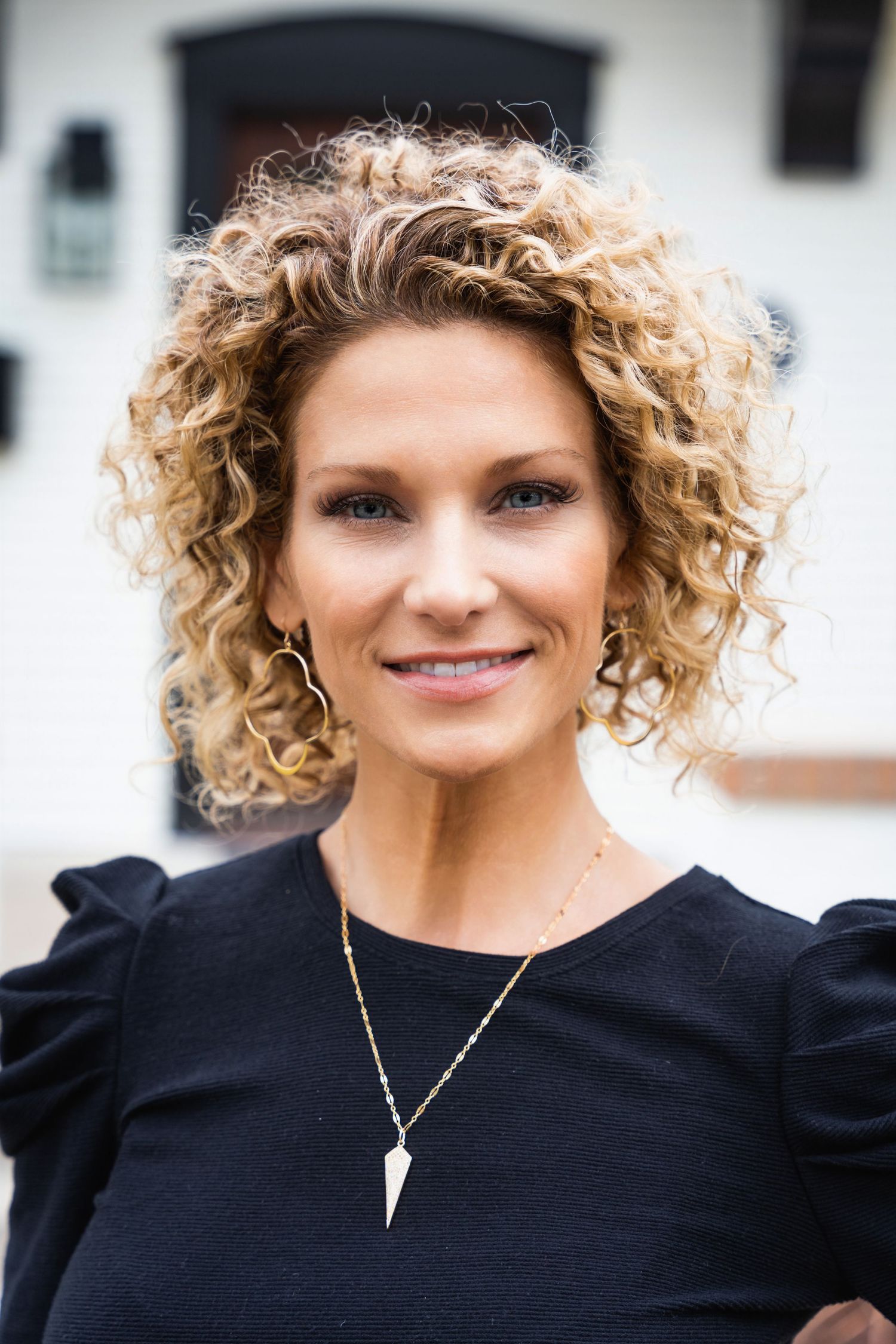Close-up portrait of person in black top with curly blonde hair and pendant necklace against white building.