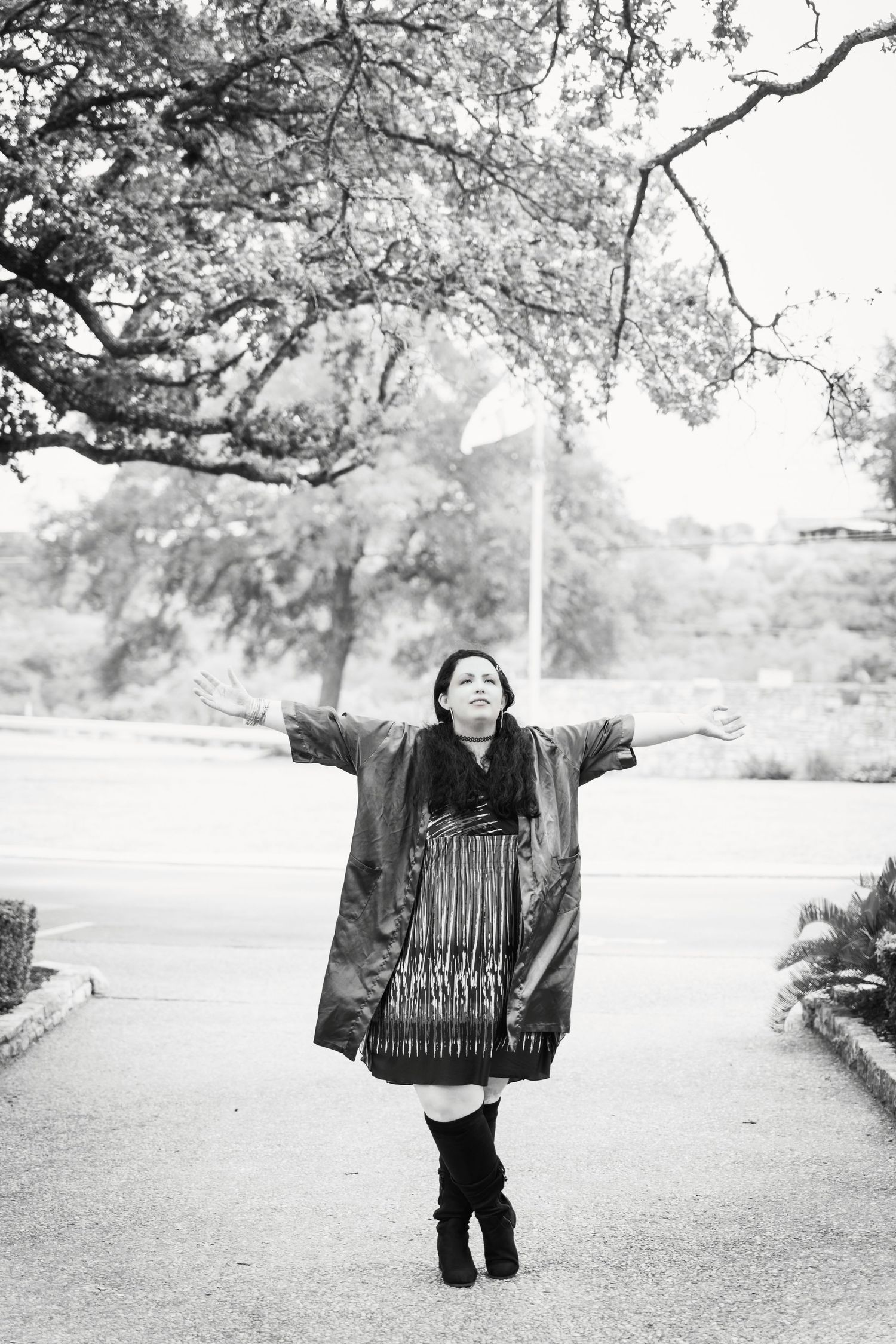 A black and white photo of someone posing with arms outstretched on a path under blooming trees.