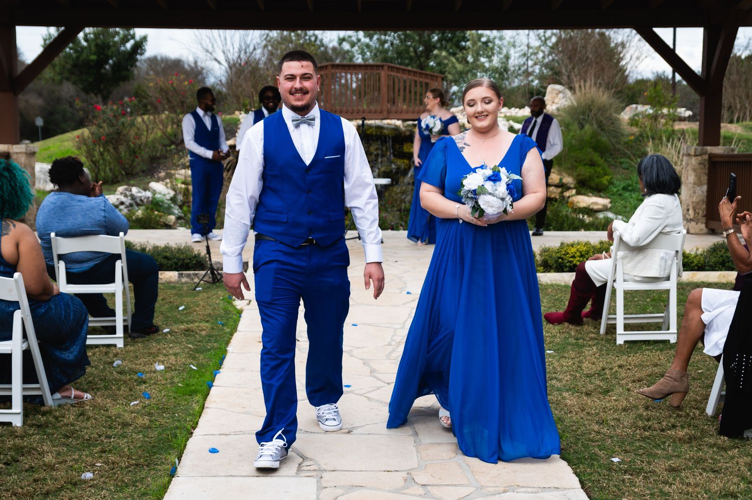 Wedding party members walk down a stone pathway at an outdoor ceremony wearing coordinated blue attire.