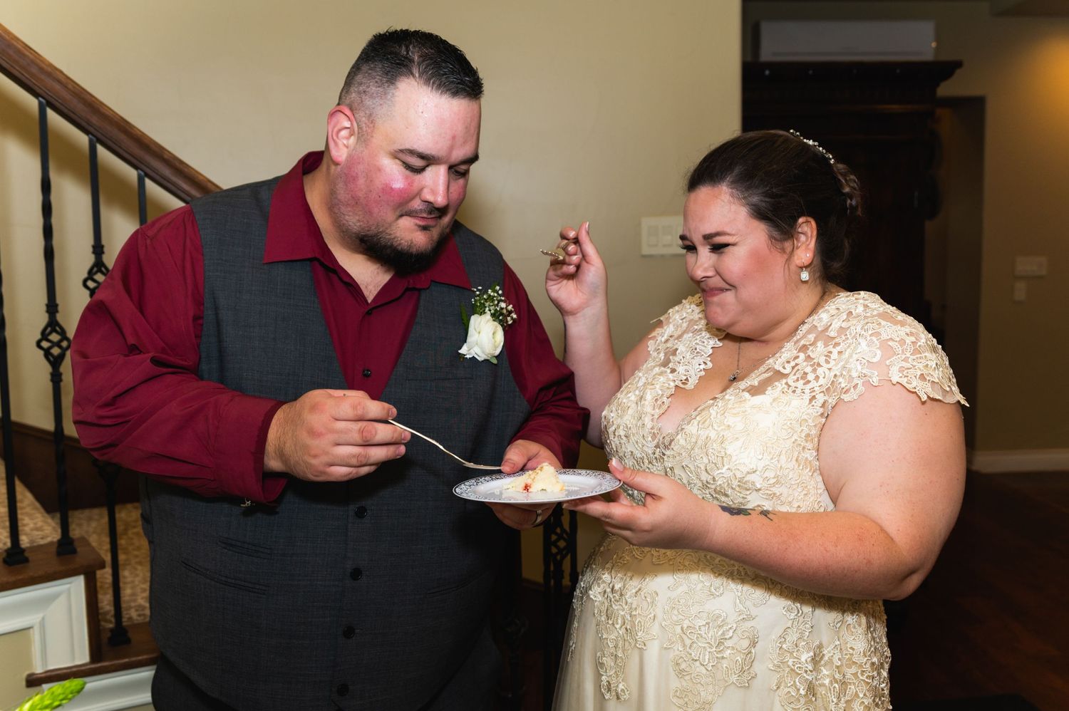 A couple shares a piece of wedding cake together during their reception near a staircase.