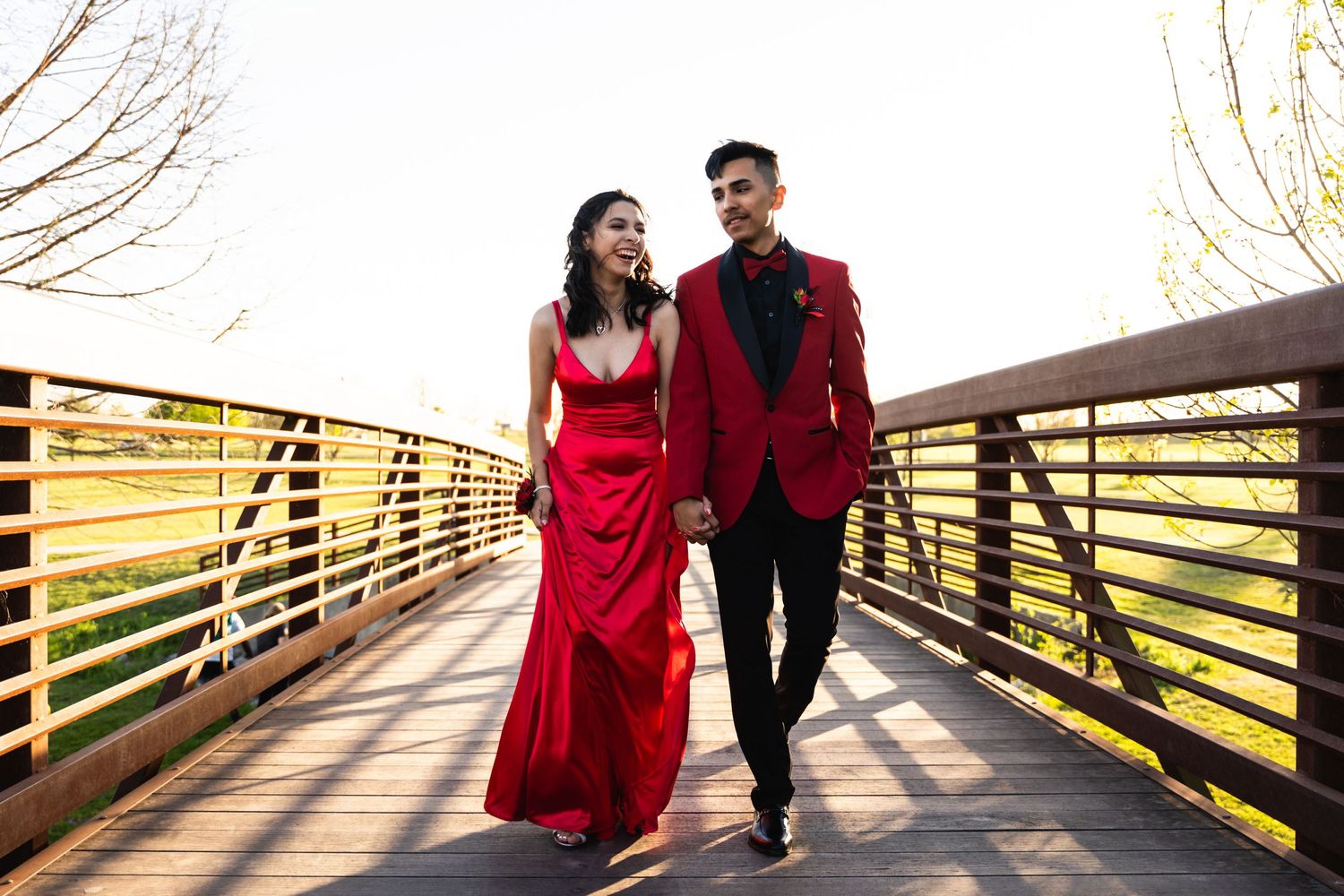 Couple in formal red attire walking together on wooden bridge during golden hour.