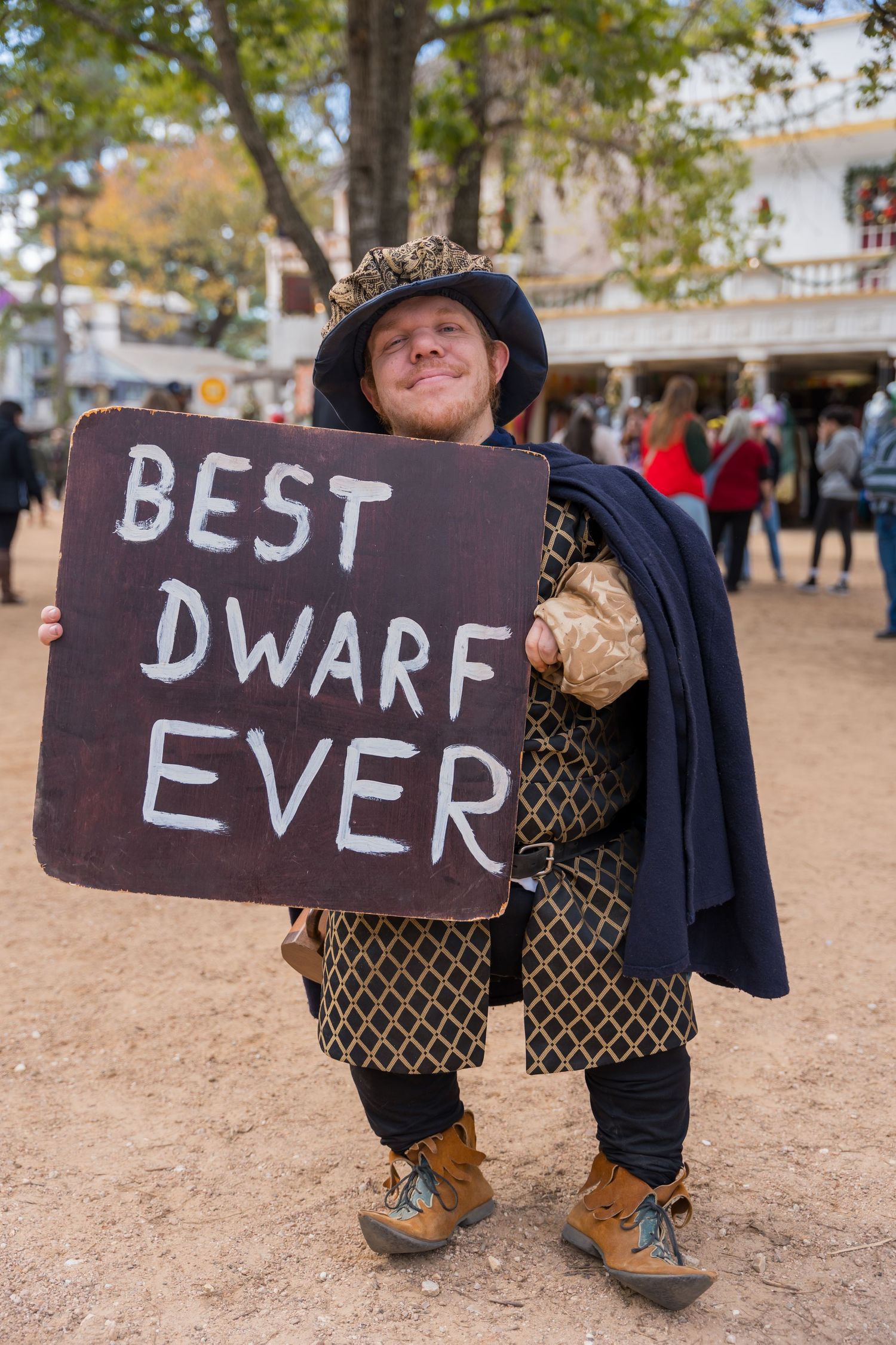 Costumed festival performer in medieval attire holding a sign reading 'BEST DWARF EVER' at a Renaissance faire.