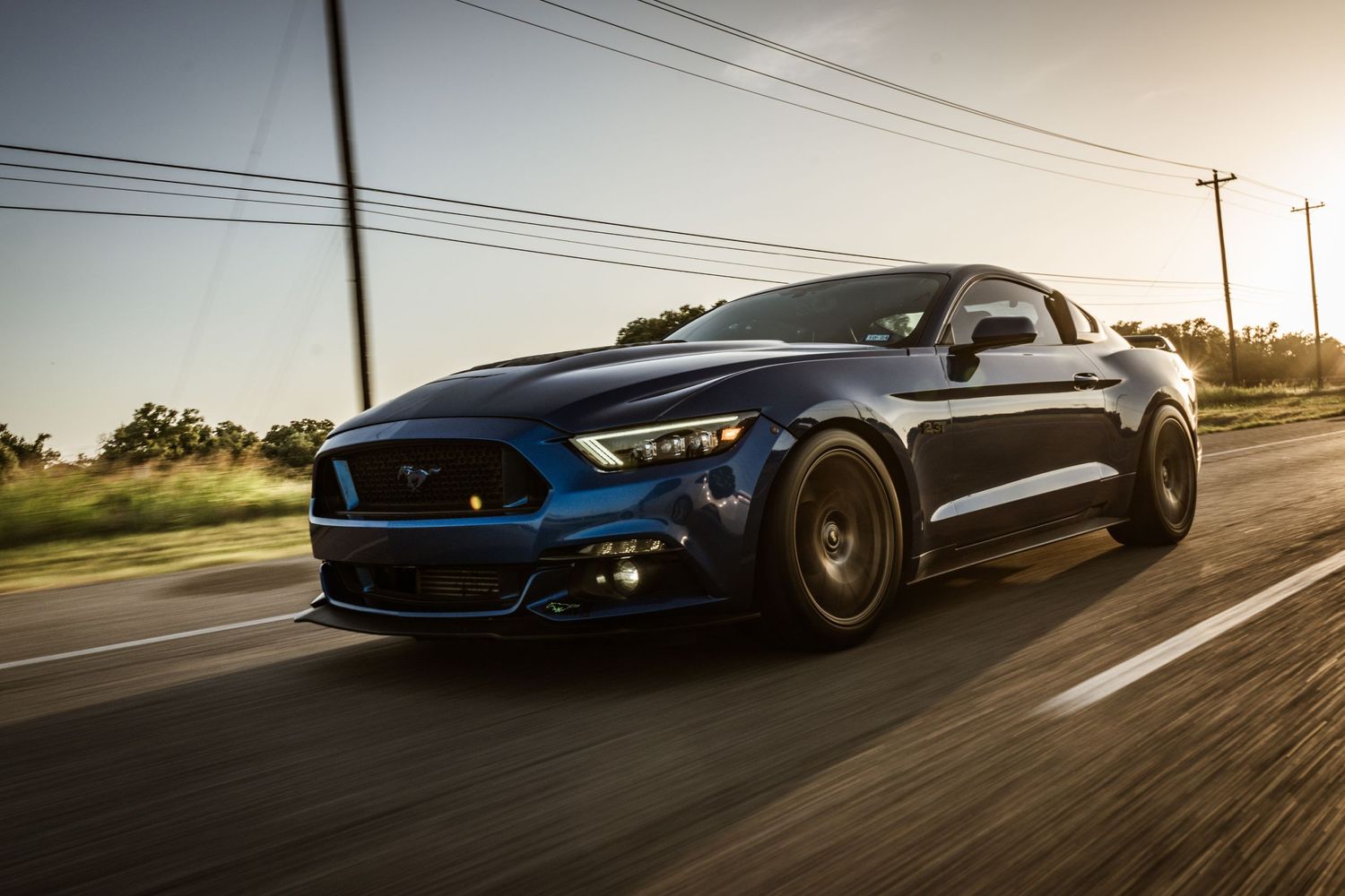 Blue Ford Mustang with aggressive body kit and black wheels driving at high speed.