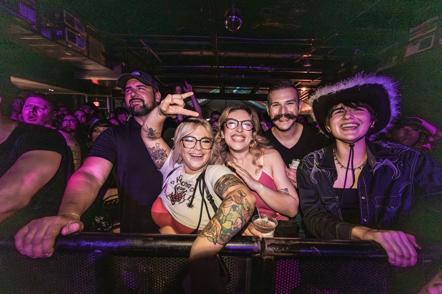 Group of concert-goers pose together at barrier with purple venue lighting.