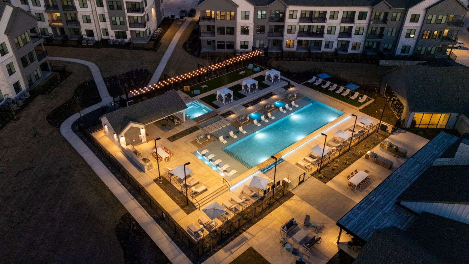 Aerial view of an illuminated luxury apartment complex pool deck with lounge chairs and palm trees at dusk.