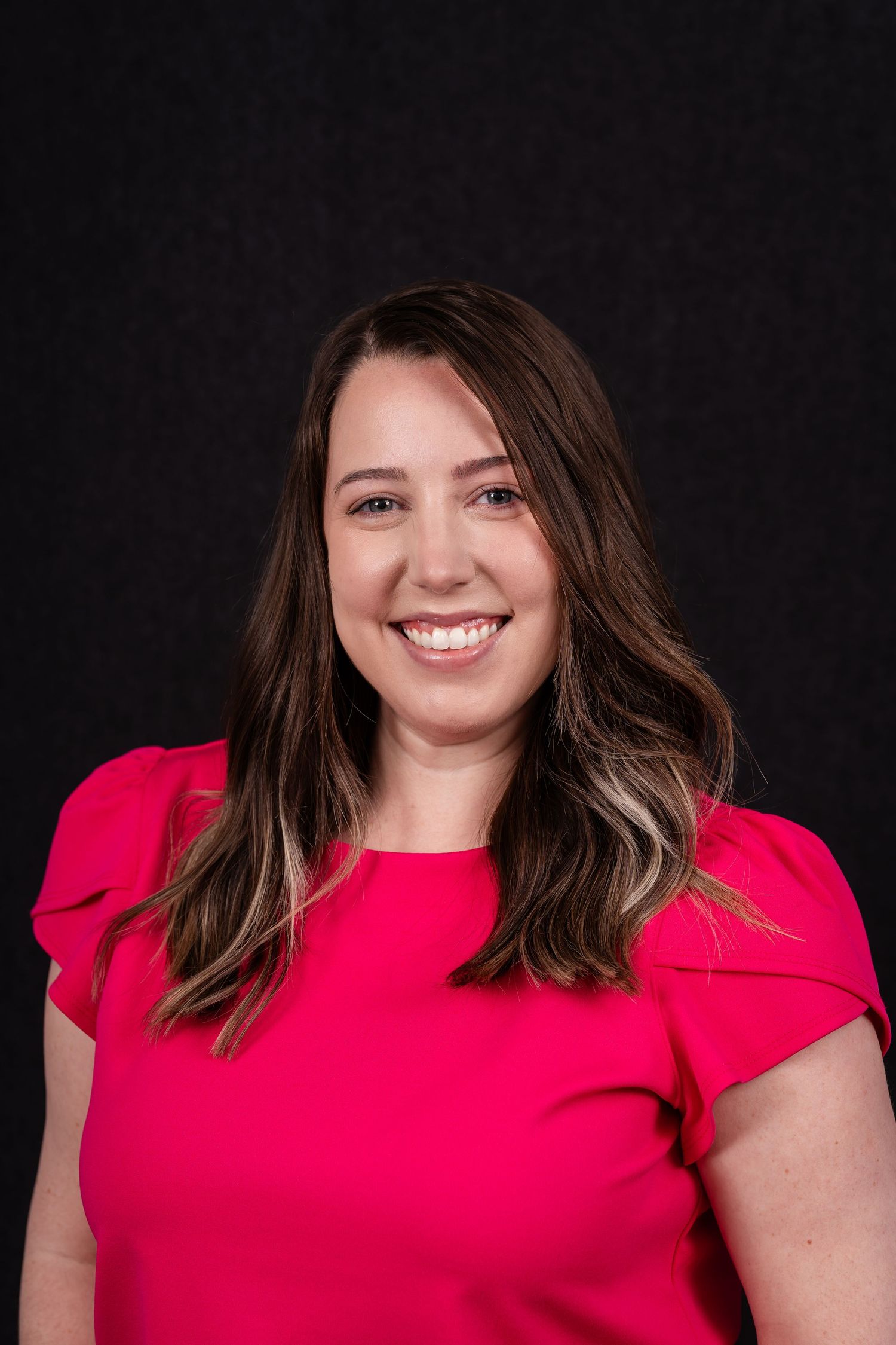 Professional headshot of a confident person in a bright red blouse smiling warmly against a dark background.