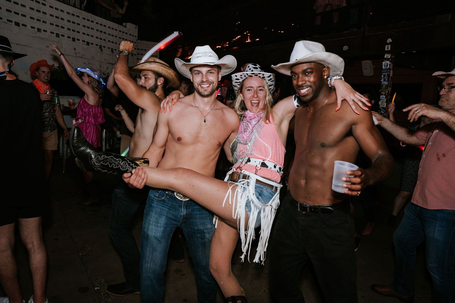 Party guests in cowboy hats and western wear celebrate energetically at an outdoor event.
