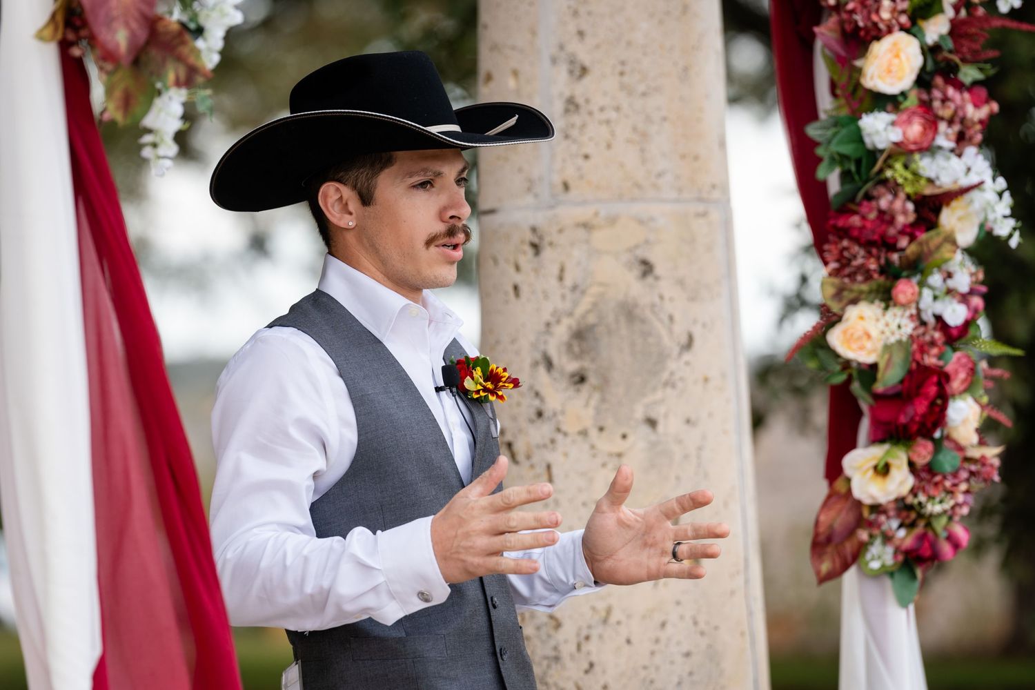A person in western attire speaks during a wedding ceremony decorated with burgundy floral arrangements.