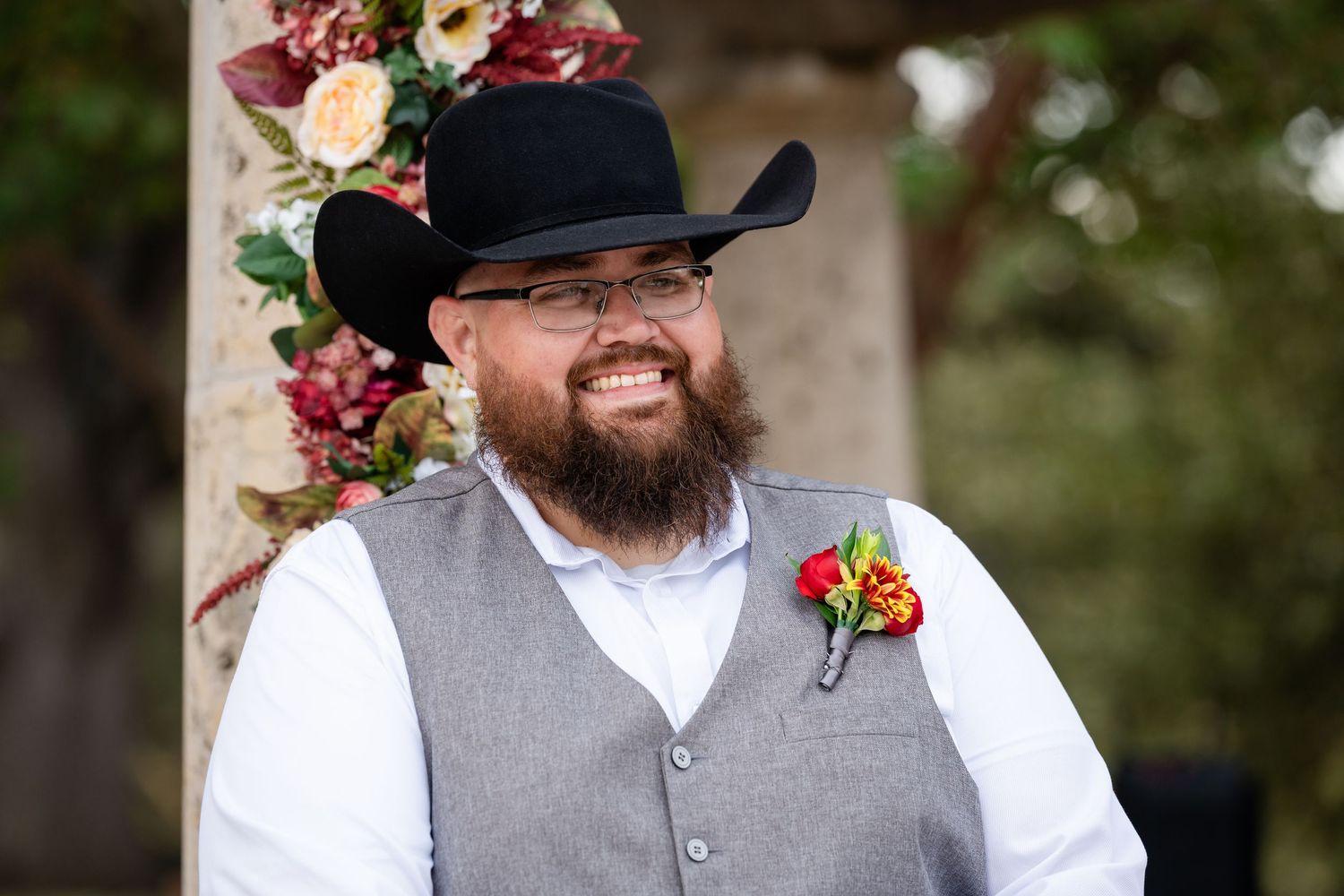 A groomsman in western attire with black cowboy hat and grey vest smiles near floral decorations.