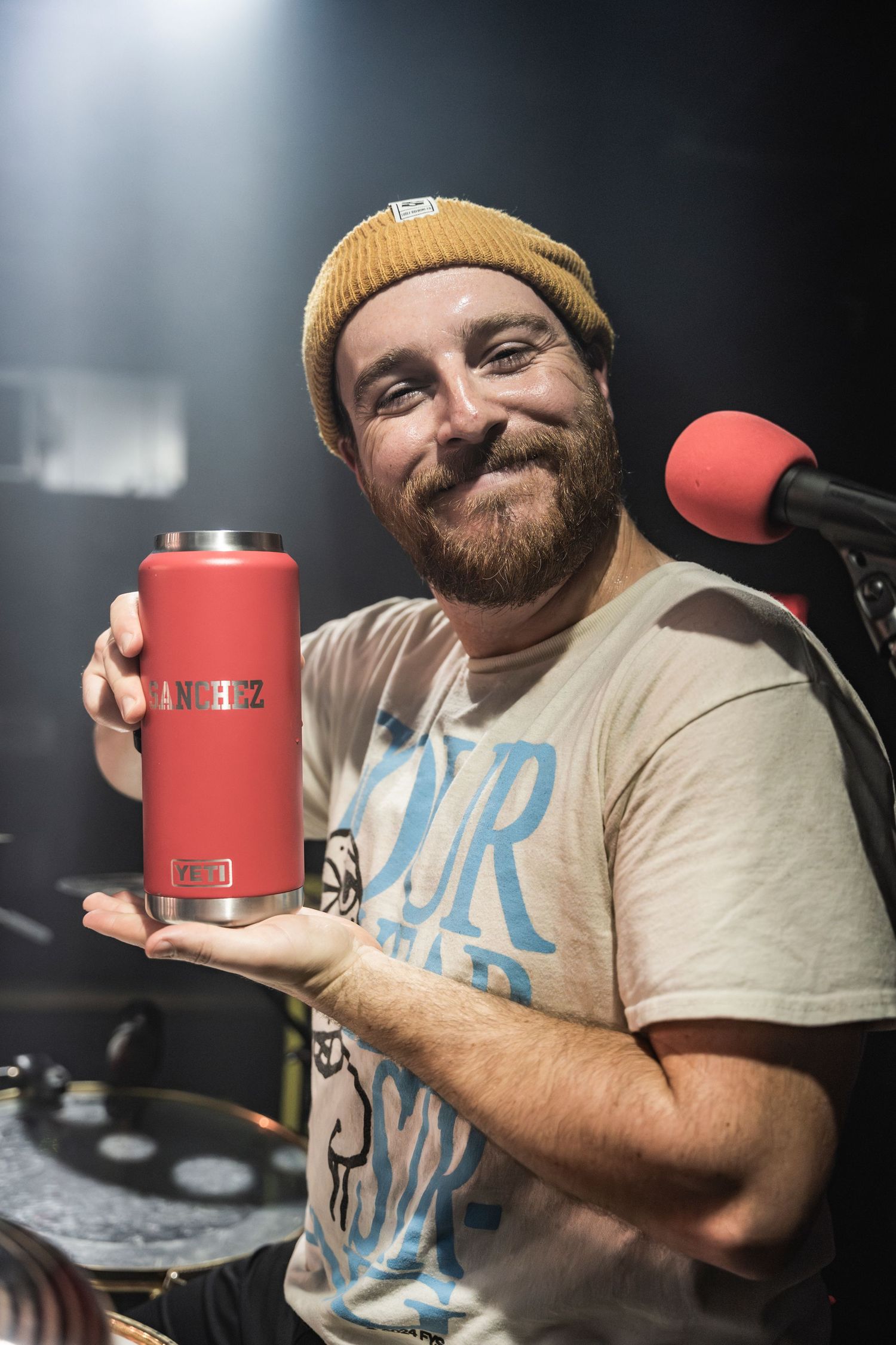 A bearded podcaster in a yellow beanie and casual t-shirt holds up a red beverage container while recording at a microphone.