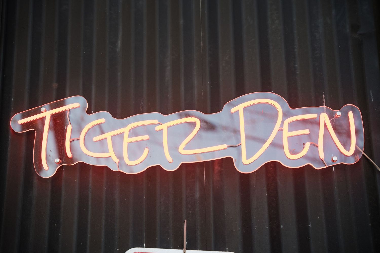 Neon sign reading 'Tiger Den' glows against a dark corrugated wall in red and white light.