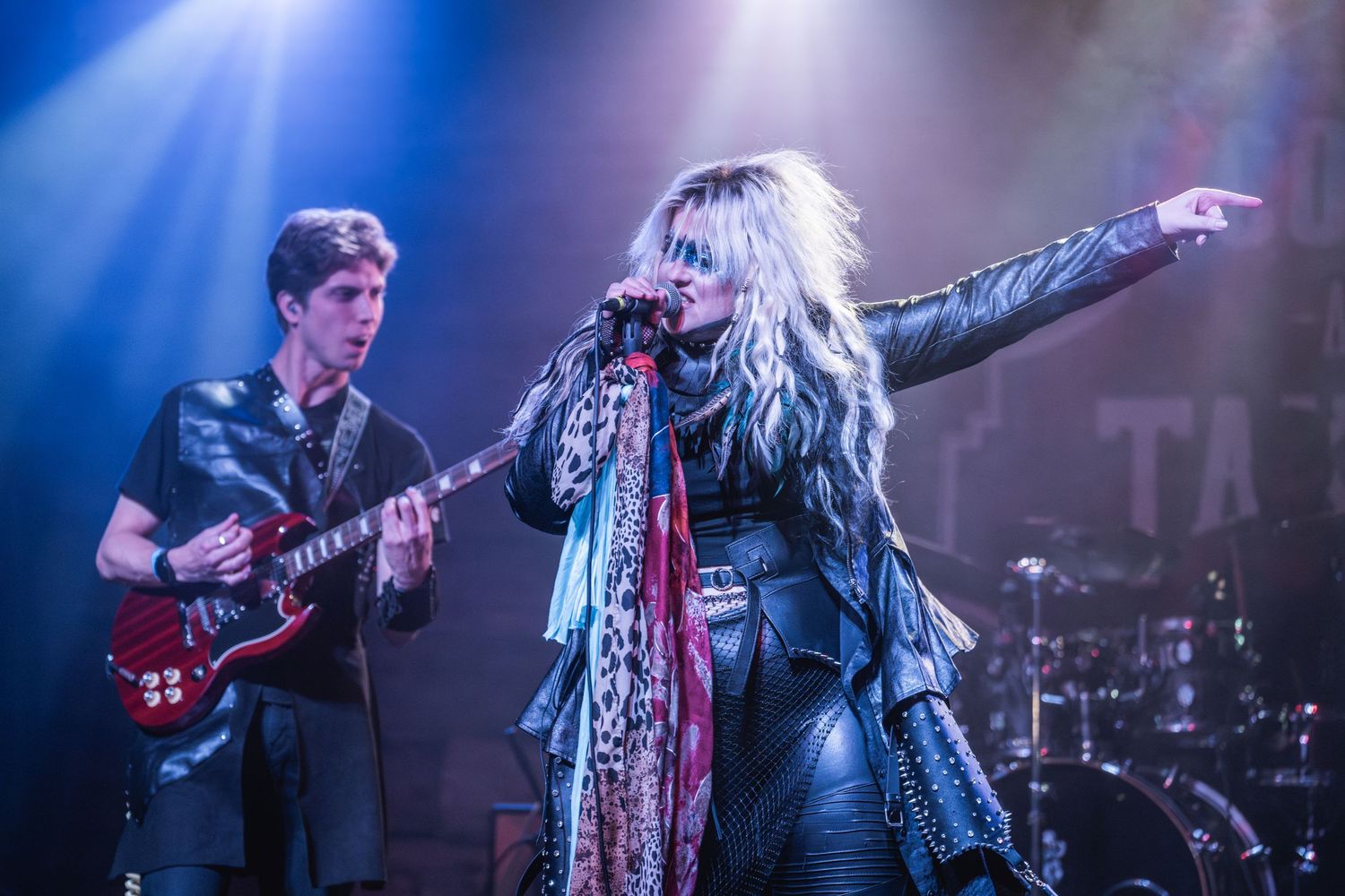 Musicians perform on stage with dramatic blue lighting while singer gestures during theatrical performance.