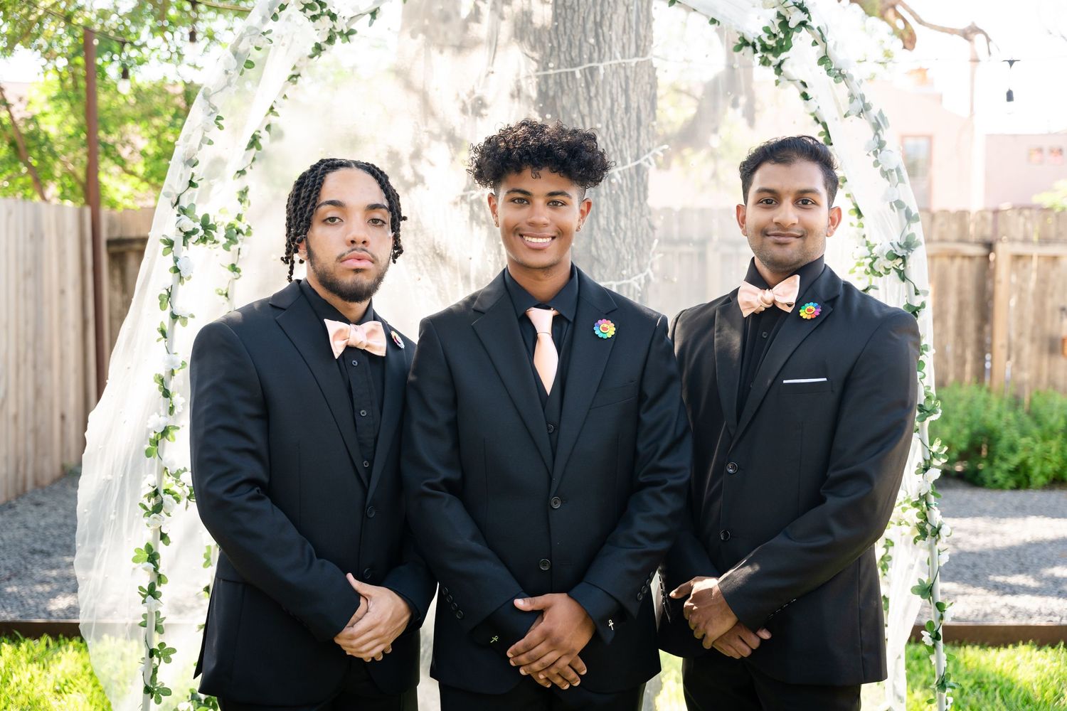 Three groomsmen in black suits and ties pose under a floral arch in a garden setting.