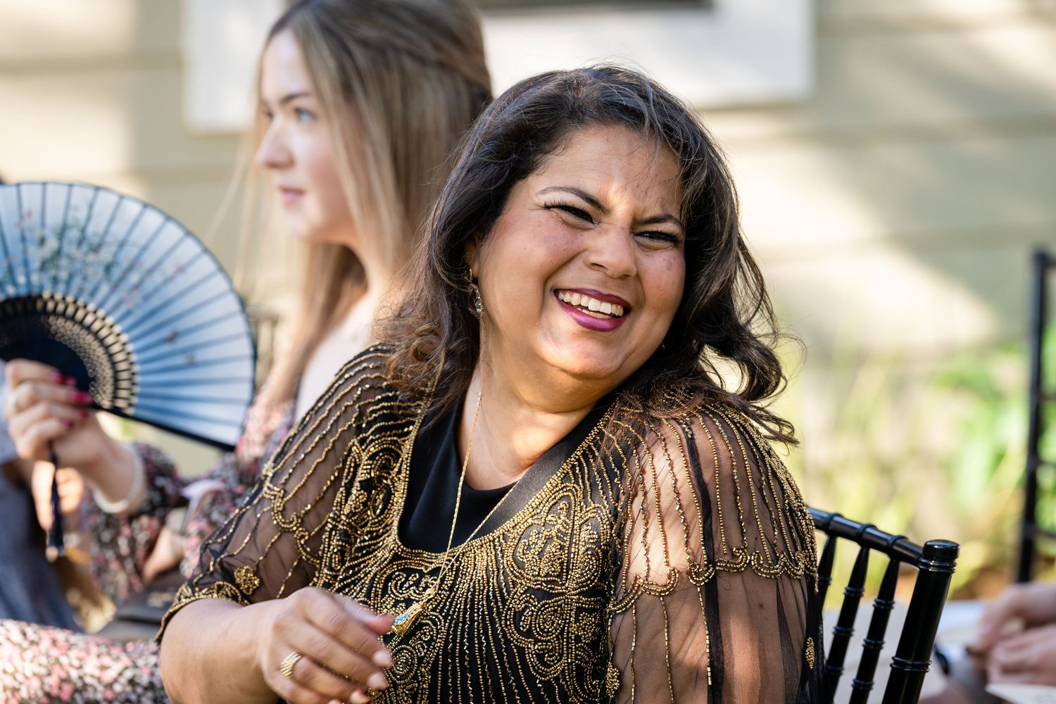A guest in a shimmering gold dress laughs joyfully at an outdoor celebration.