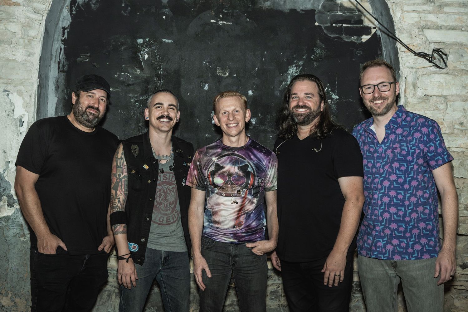 Band members stand together against rustic brick wall background.