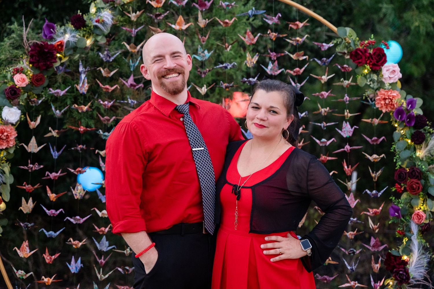 Couple poses in red and black attire in front of decorative paper crane backdrop.