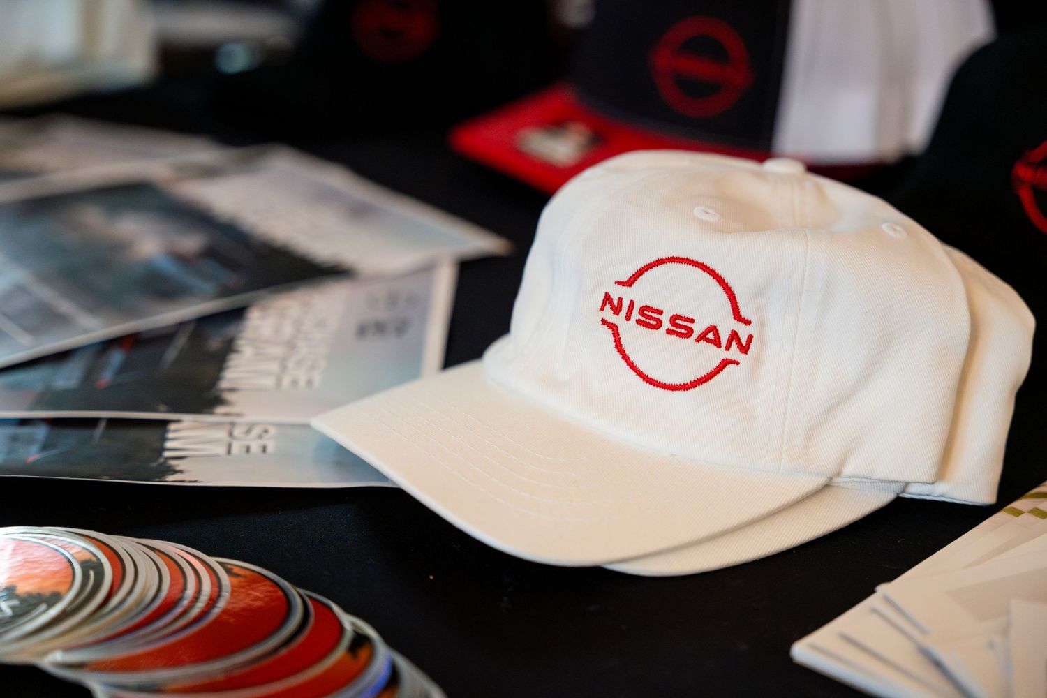 A white baseball cap with the red Nissan logo sits on a table next to scattered photographs.