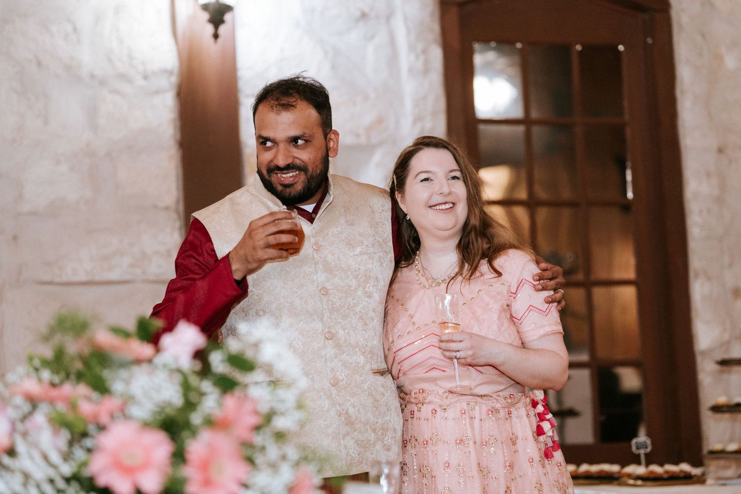 A couple celebrates at a wedding reception wearing pink and red traditional attire with floral decorations.