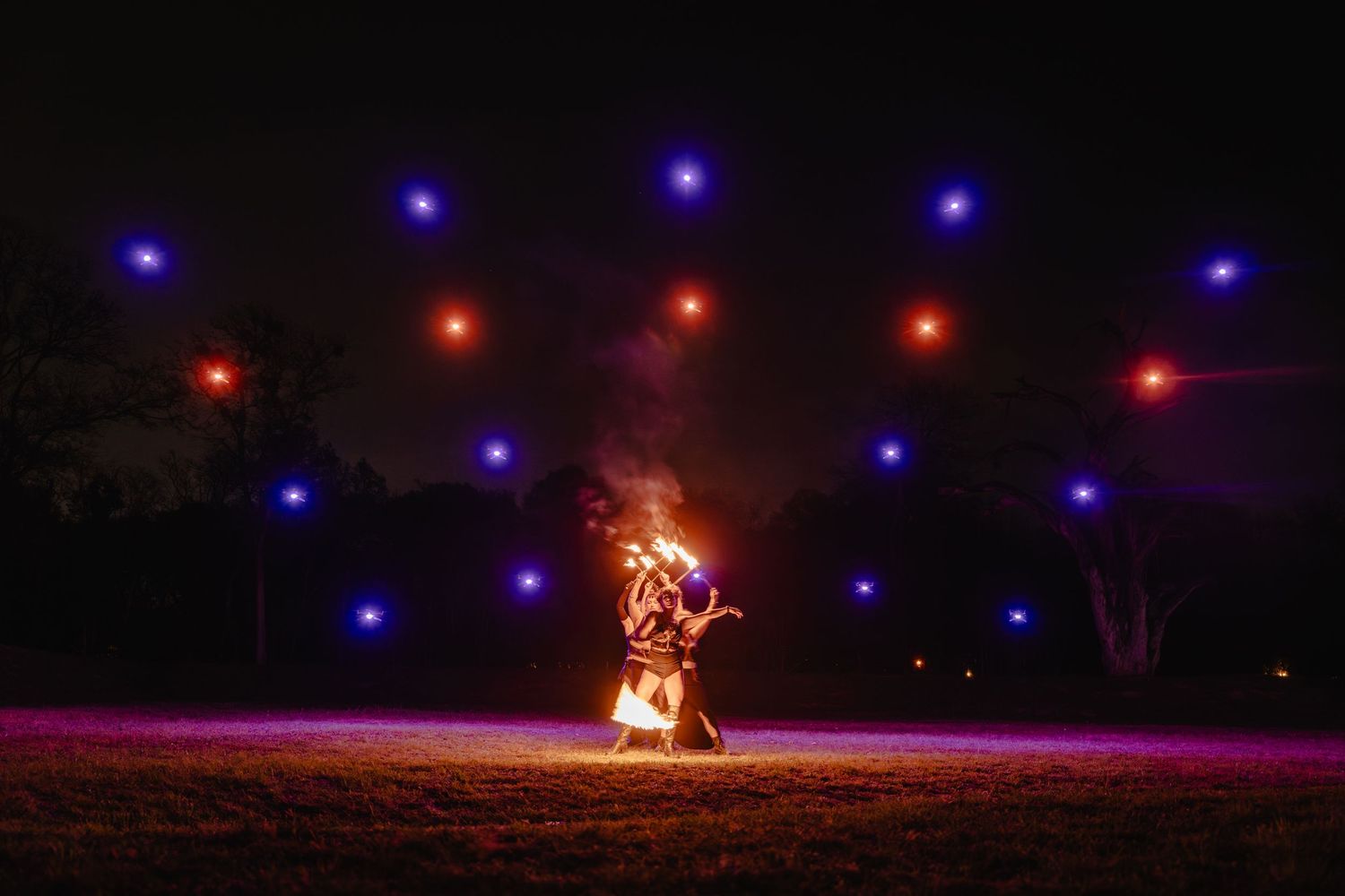 Fire performer spinning flames at night with colorful blue and red lights arranged in an arc pattern overhead outdoors.