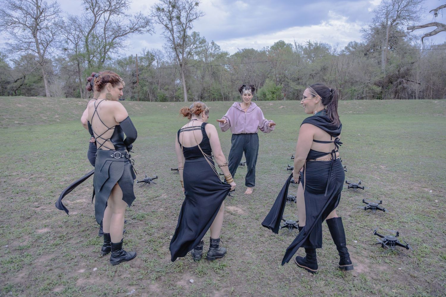 Four women in dark fantasy costumes gather in a grassy field with drones on the ground during an outdoor photoshoot.