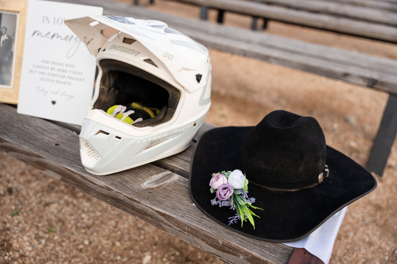 White motocross helmet and black cowboy hat with floral corsage displayed on bench at memorial tribute.