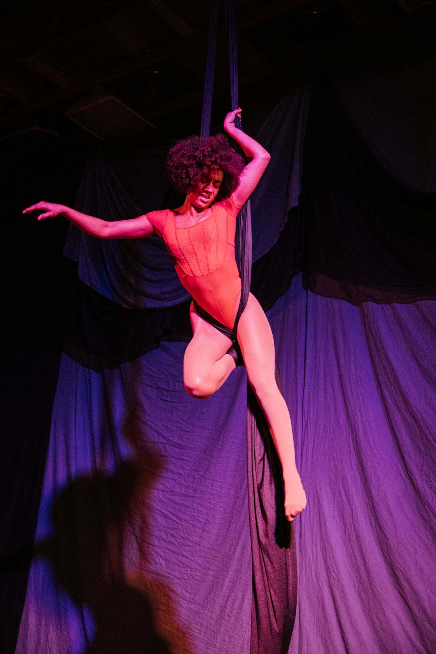 Aerial silk performer in red costume suspended mid-air against dramatic purple and black draped fabric backdrop.