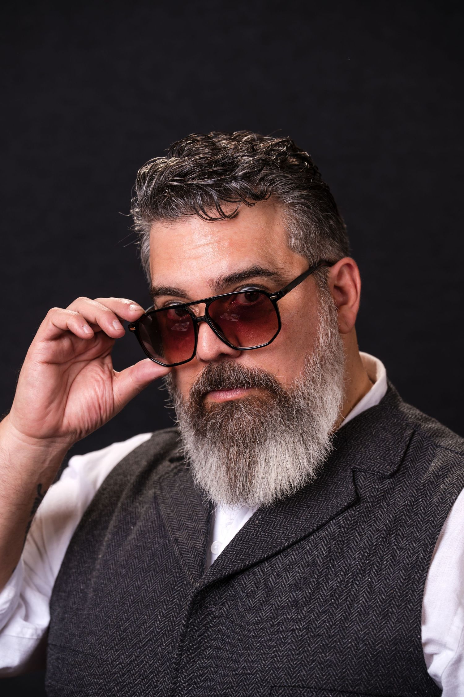 A bearded person in sunglasses and a gray suit jacket poses in a series of four similar headshots against a dark background.