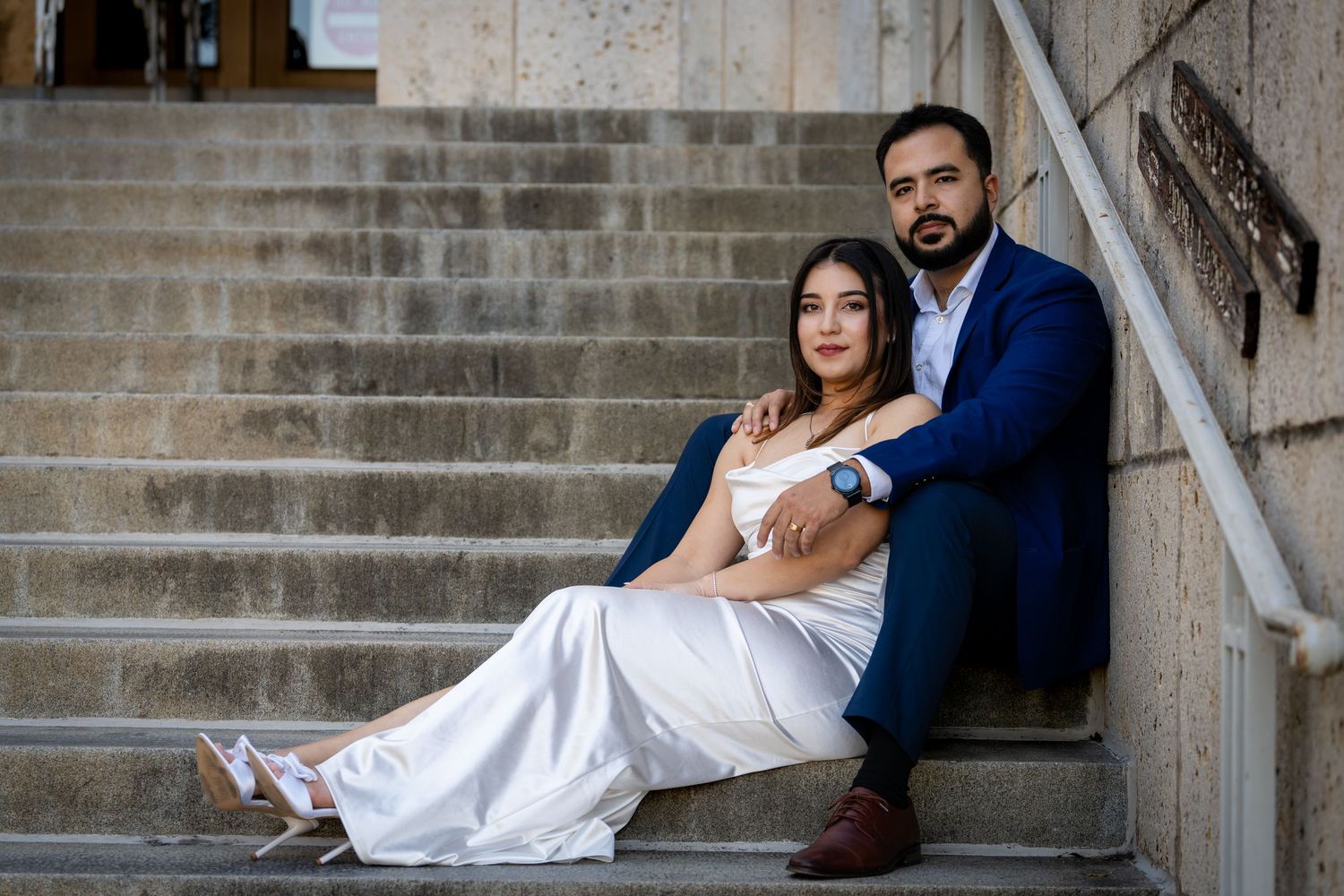 A couple in formal attire poses romantically on stone steps for wedding photos at dusk.