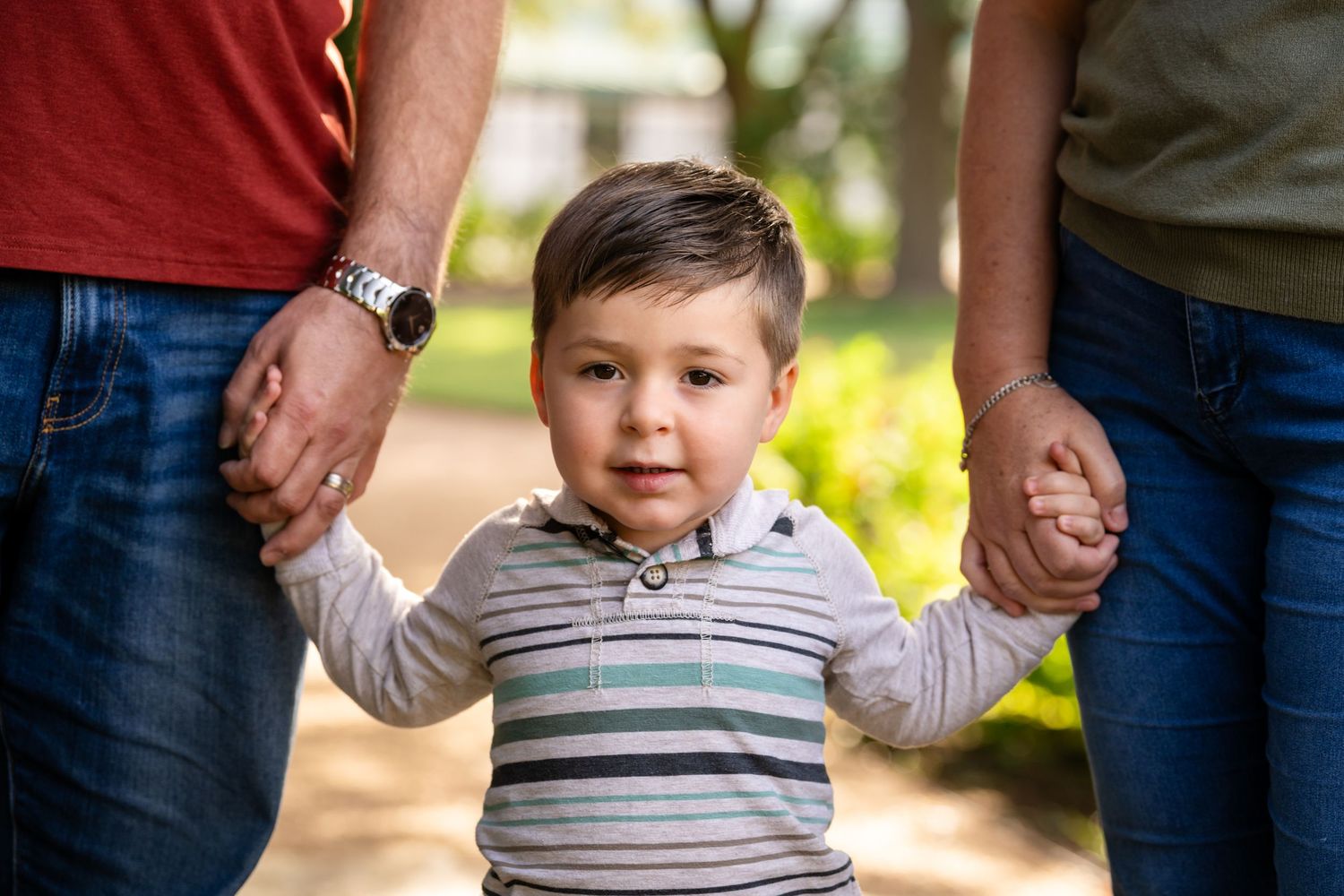 A toddler in a striped shirt holds hands with adults while walking outdoors in a park on a sunny day.