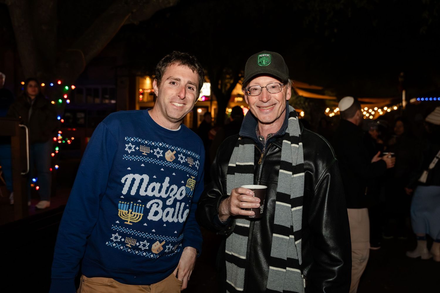 Two people at a nighttime event, one wearing a holiday sweater and the other in a striped scarf and baseball cap.