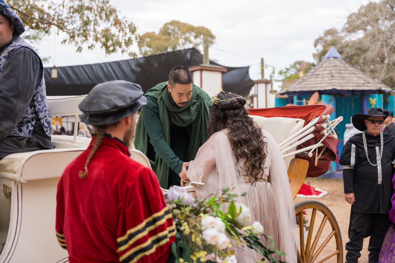 People in period costumes gather at an outdoor colonial reenactment event with rustic buildings in the background.