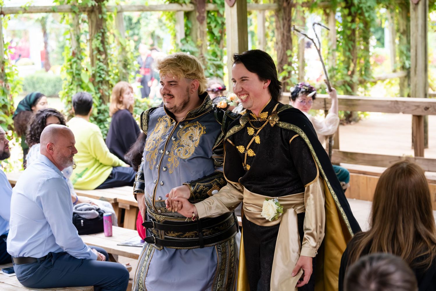 People gather and socialize at a covered outdoor venue with greenery and decorative elements visible in the background.