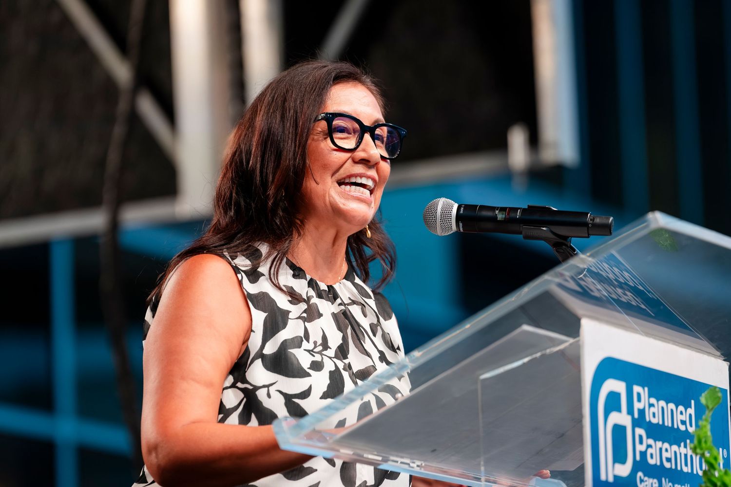 Speaker stands at podium wearing black and white patterned dress and glasses during presentation.