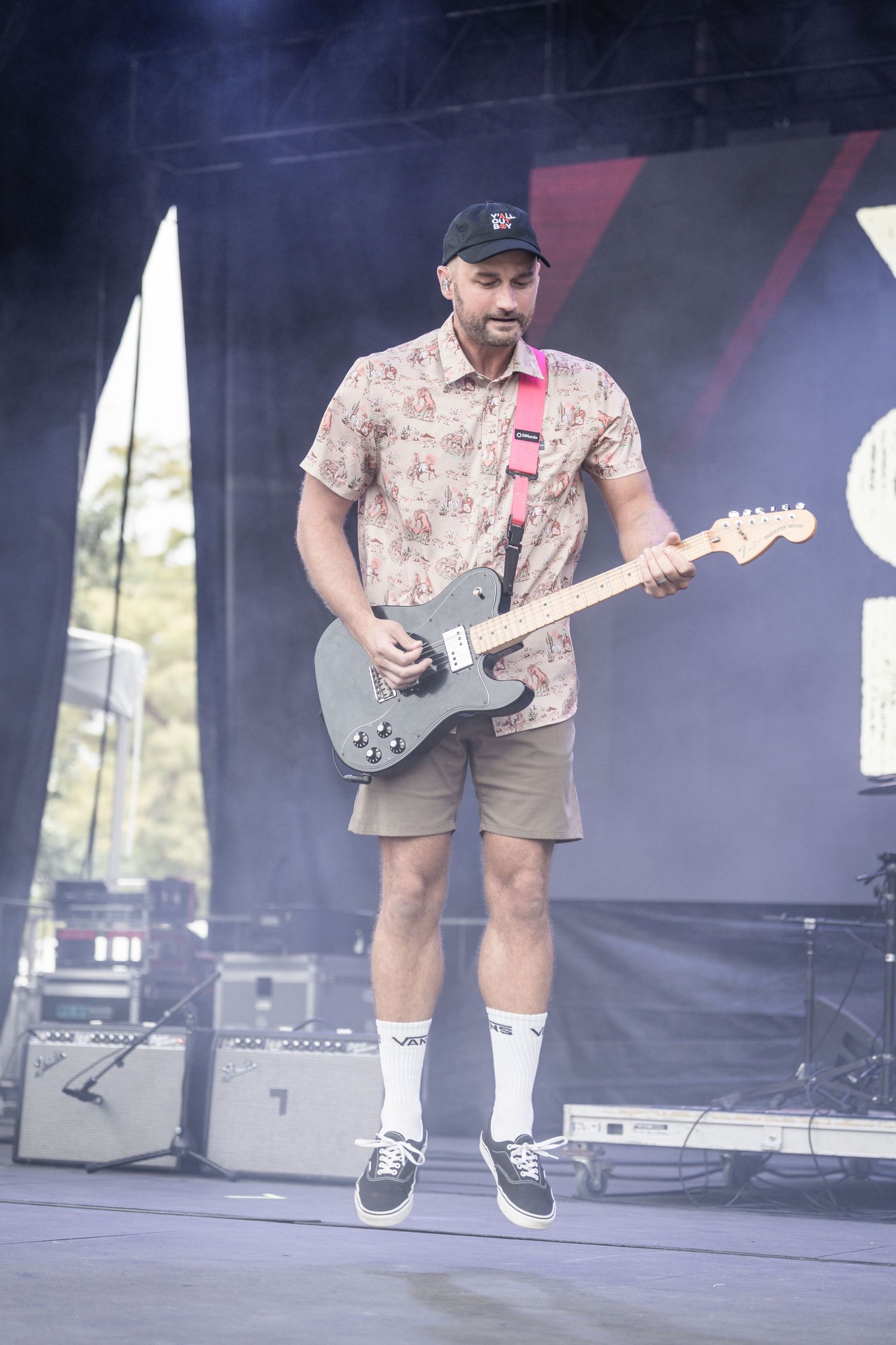 Musician performs on stage wearing floral shirt and shorts while jumping with electric guitar at outdoor concert.