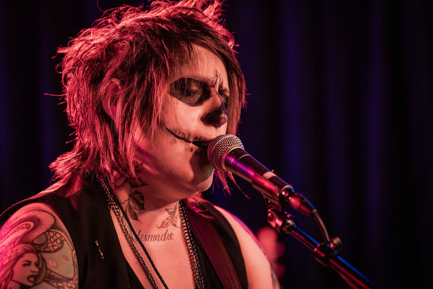 A performer with spiky hair and tattoos sings into a microphone on stage under dramatic red lighting.