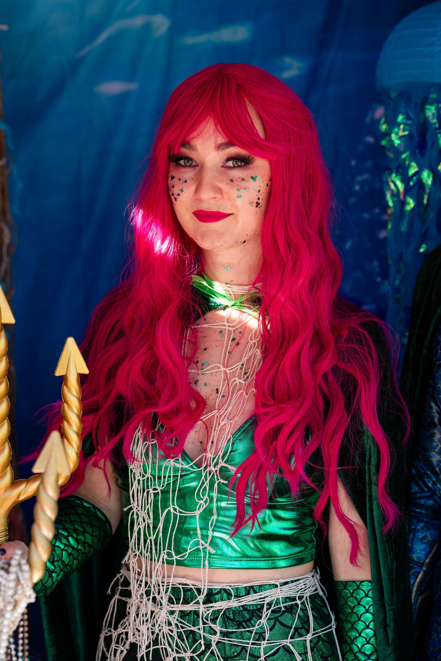 A figure with vibrant red hair poses in a metallic green costume against a dark blue background.