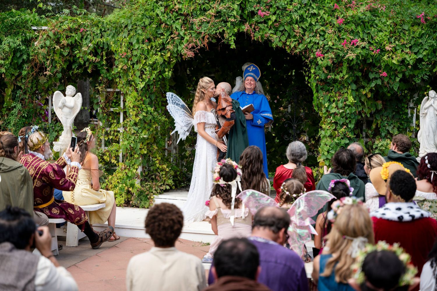 Outdoor wedding ceremony in a lush garden setting with guests seated on white chairs watching the celebration.