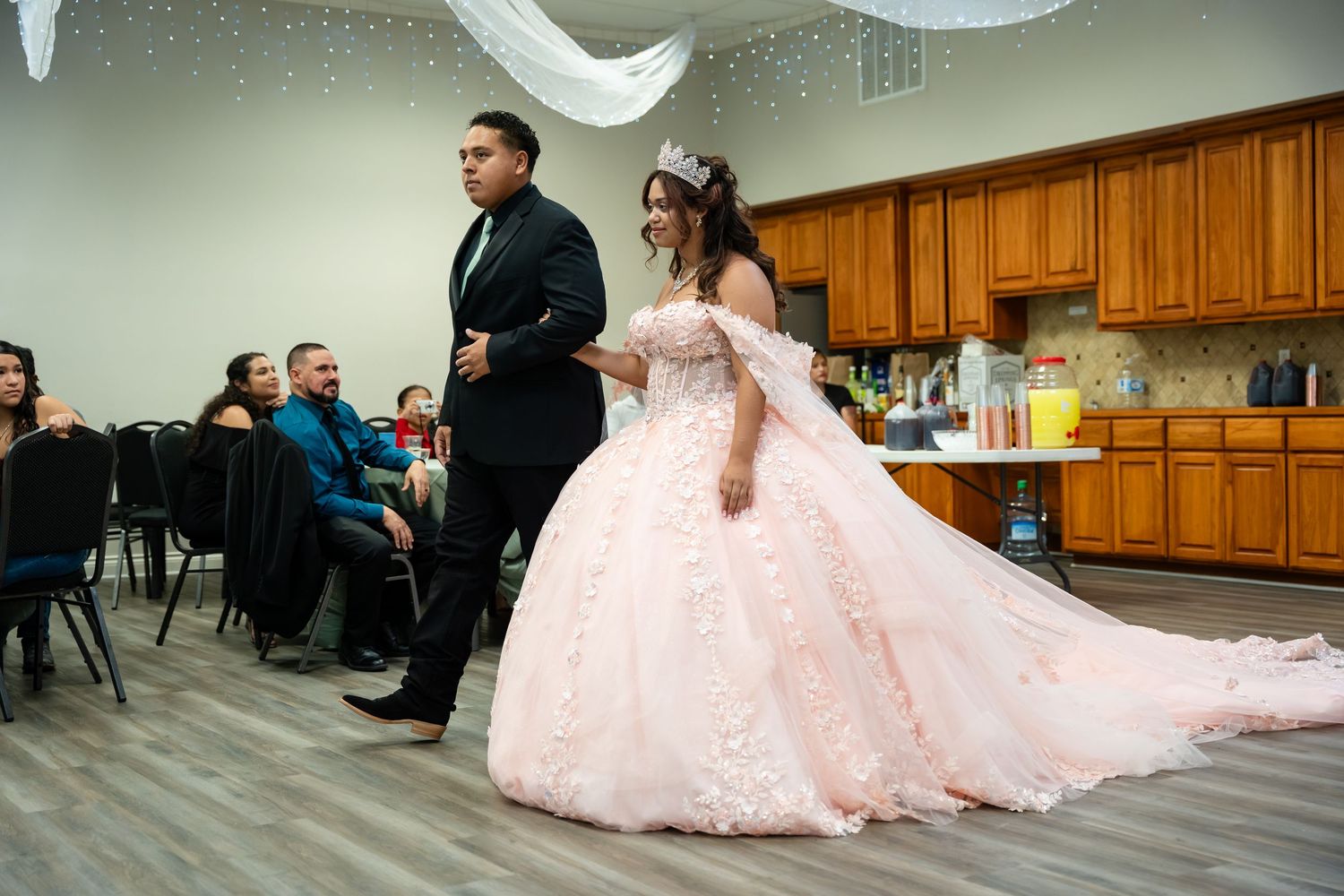 A young woman in a pink quinceañera ball gown with a tiara walks with a young man in a suit at her celebration.