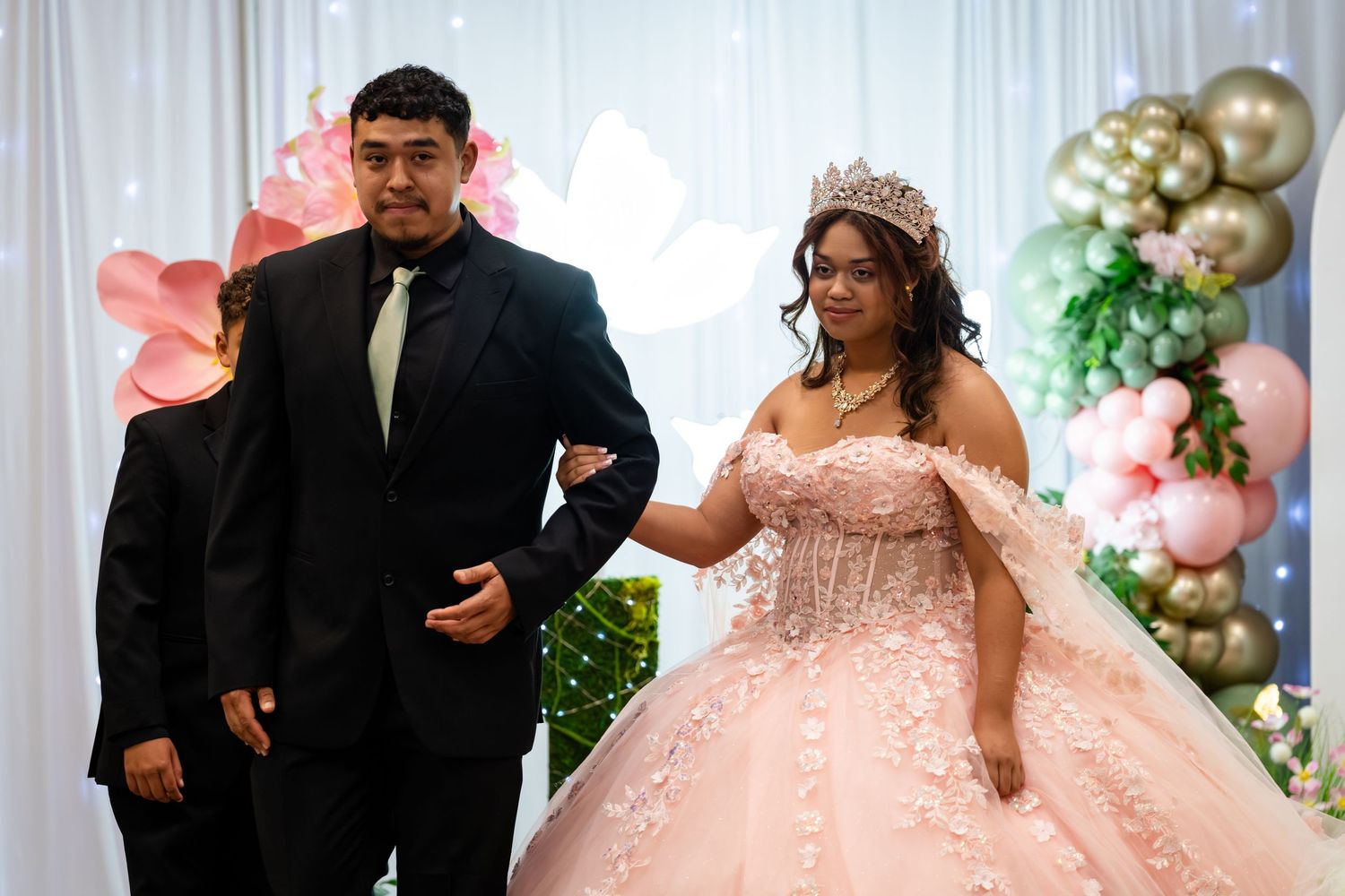 Young woman in pink quinceañera ball gown with tiara walks arm-in-arm with escort in black suit at celebration.