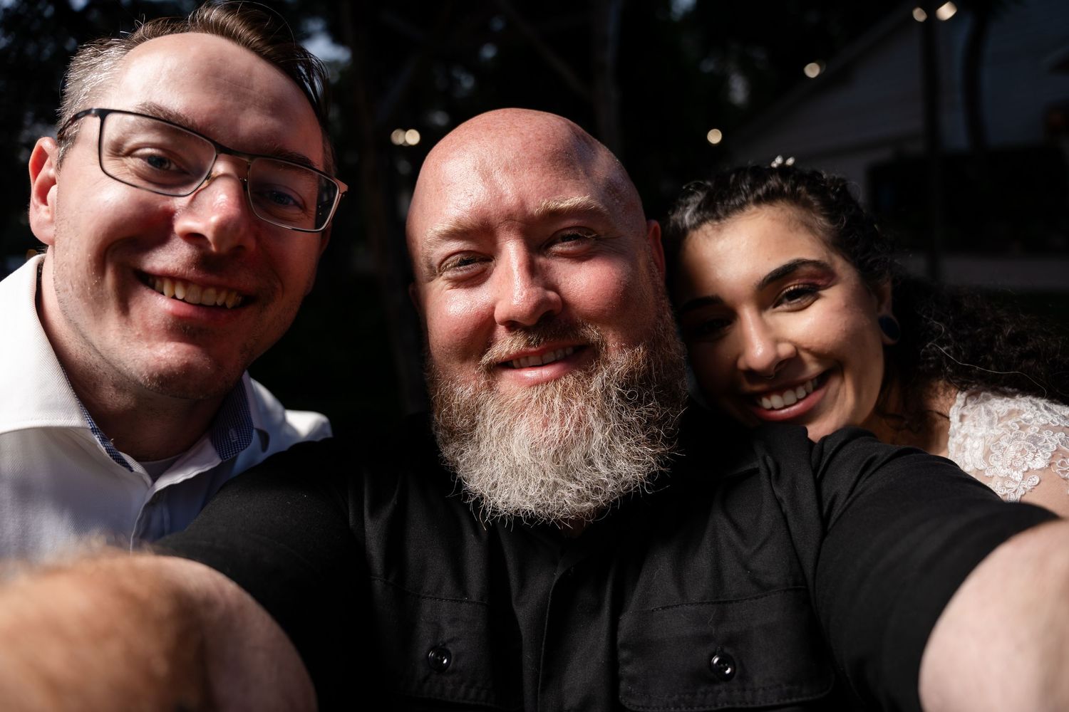 Three smiling people taking a selfie outdoors, including a bearded man in black and a woman with dark hair.
