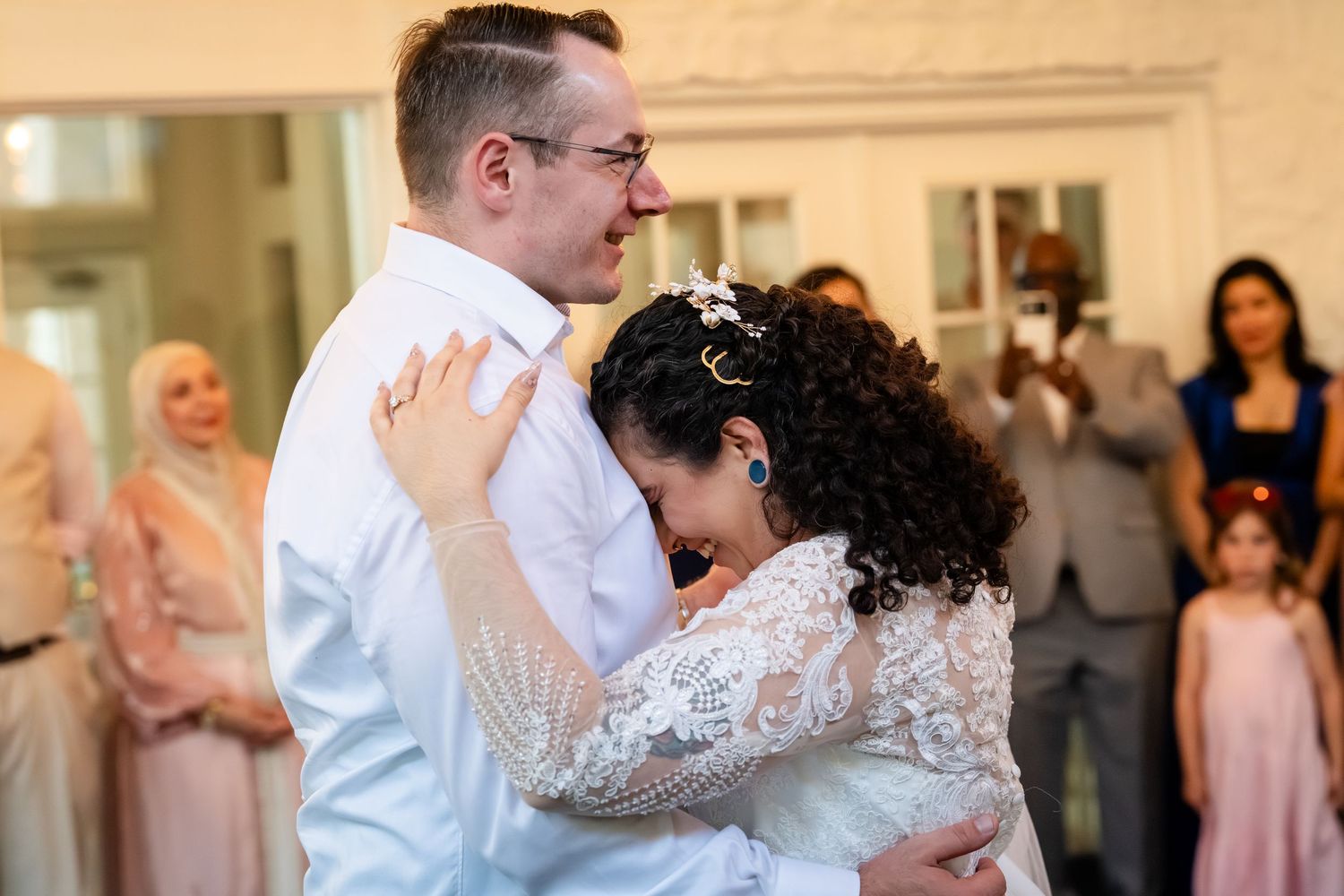 Bride and groom share emotional first dance embrace at wedding reception with guests watching in background.