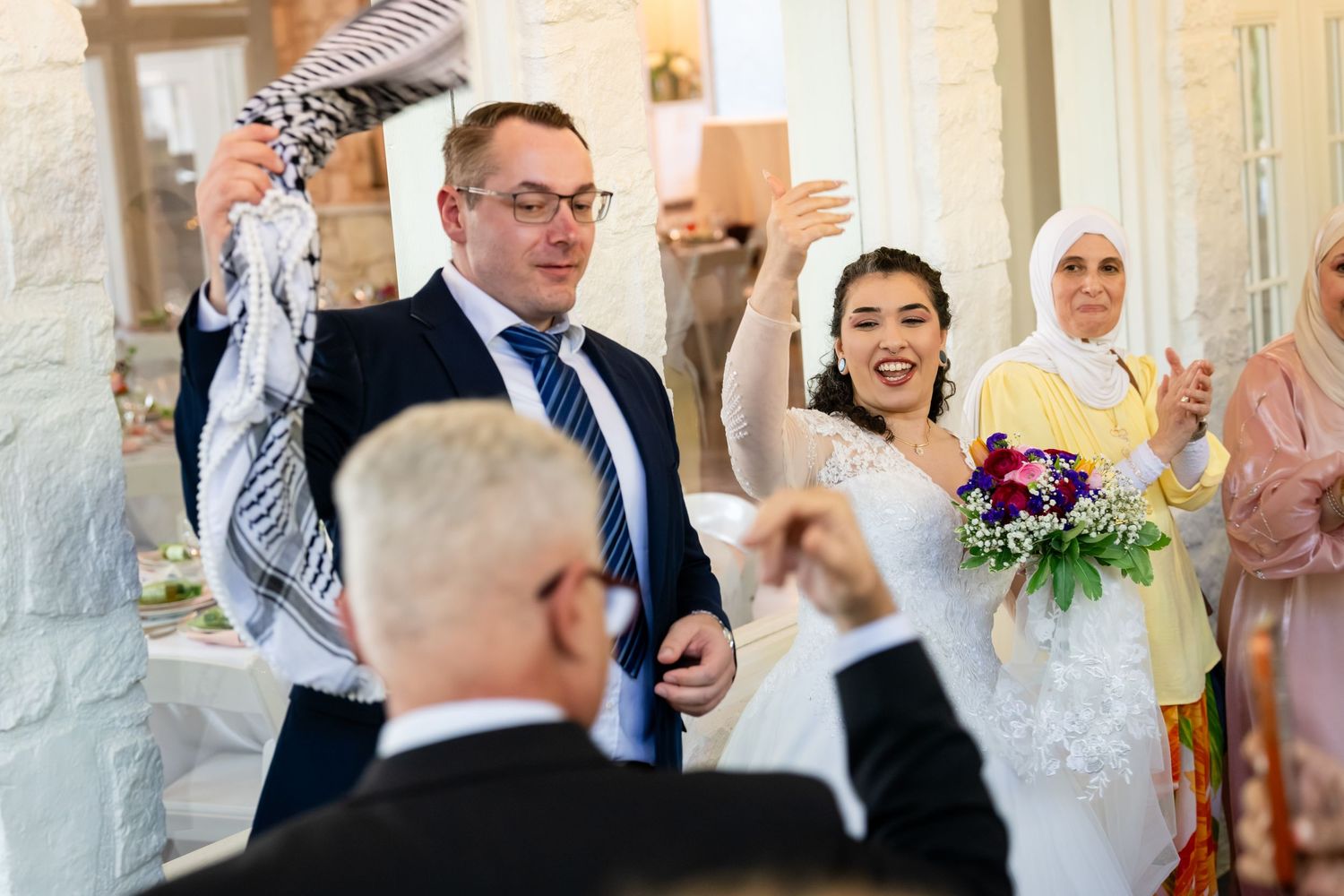Joyful wedding couple celebrating with guests, bride holding colorful bouquet in elegant venue.