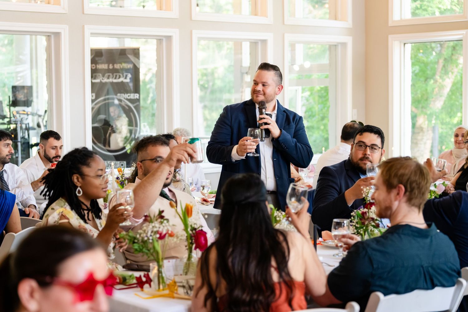 Wedding guests raise glasses during a toast speech at an elegant indoor reception venue with large windows.
